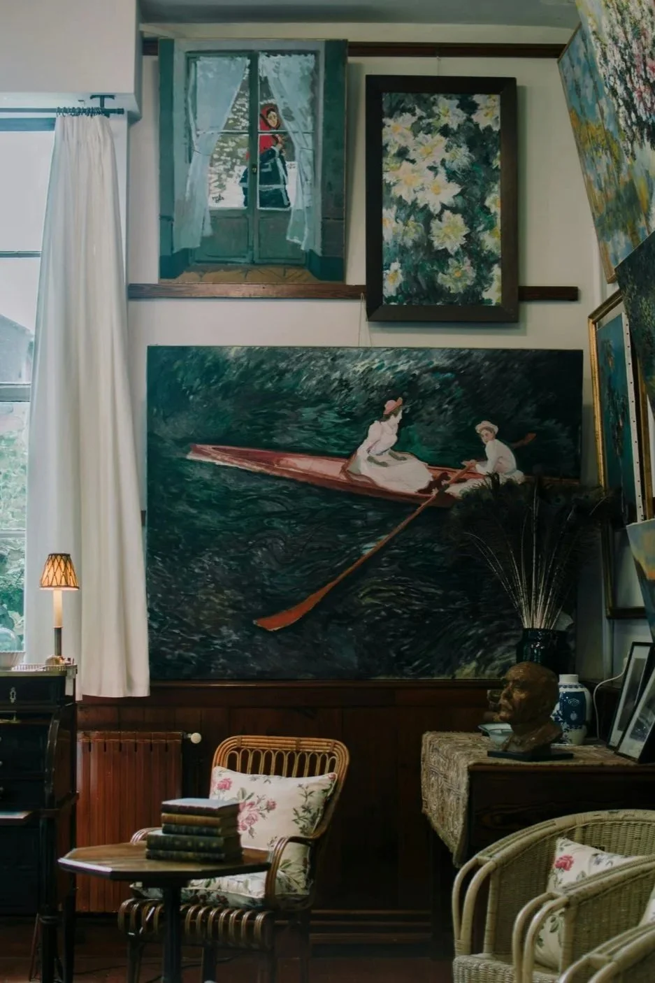 A cozy interior with various paintings on the walls, including one of two women rowing a boat. The room has a mix of vintage and artistic decor, with a small side table, wicker chair, and framed photographs.