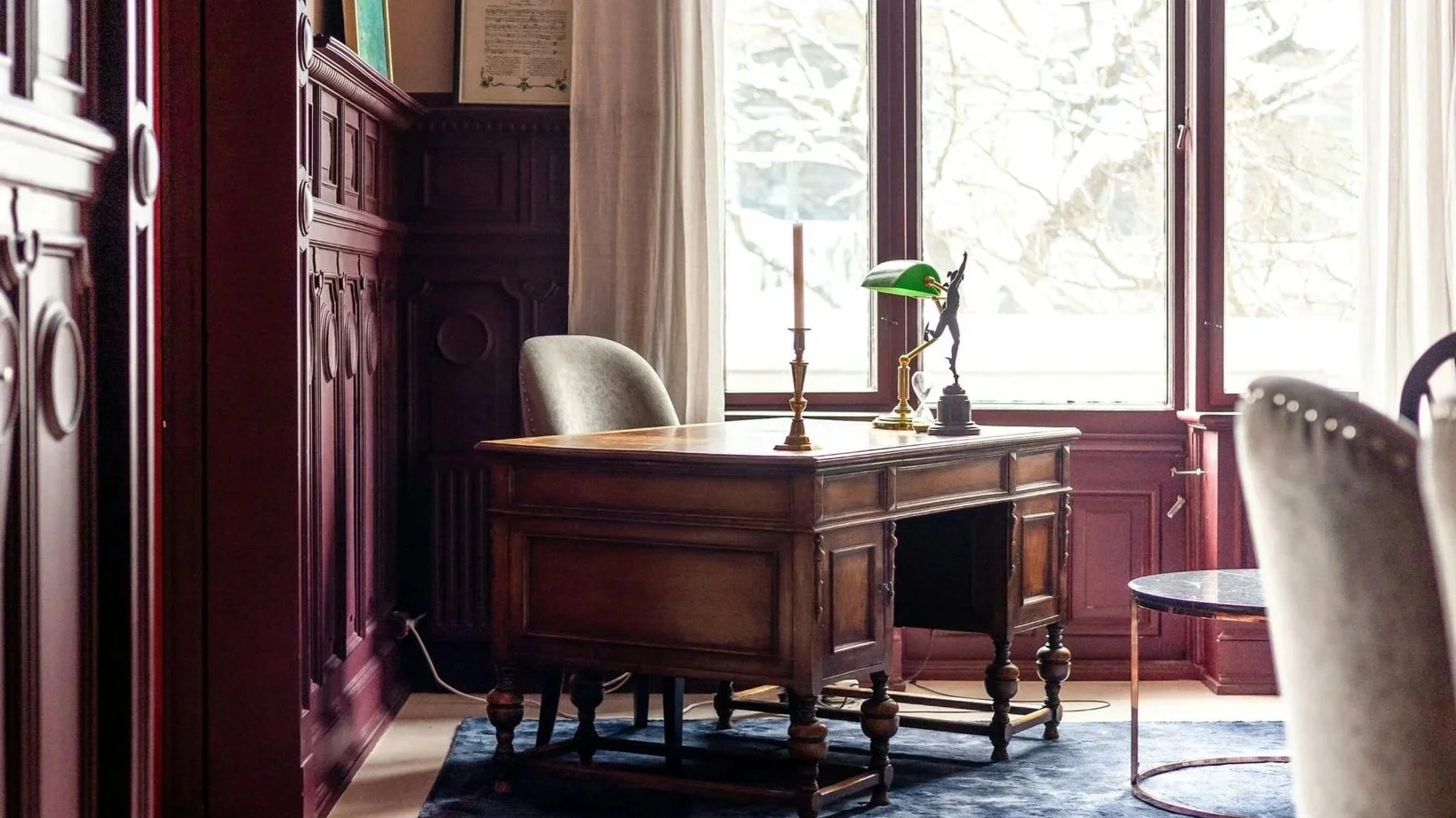 A classic wooden desk with a green banker’s lamp, a brass candlestick with a candle, and a small statue of a dancer, positioned in front of large windows with sheer curtains, in a richly paneled room.