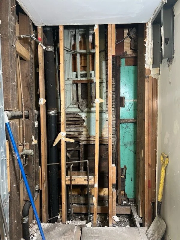 Interior of a construction or demolition site revealing exposed wooden studs, pipes, and an old wall with chipped paint.