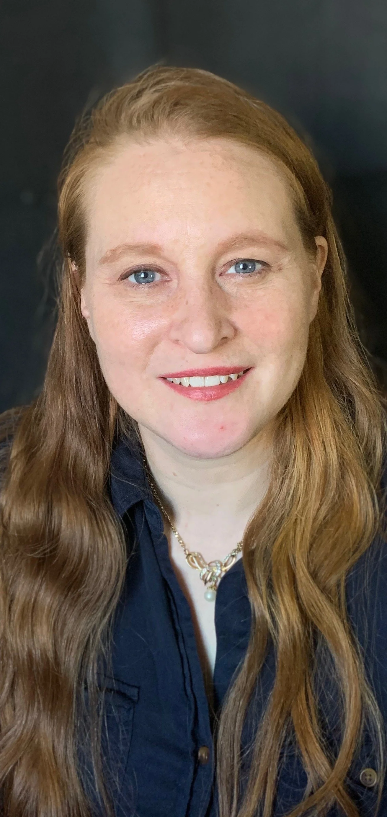 Smiling person with red hair sitting on a sofa or chair, wearing a navy blue top.