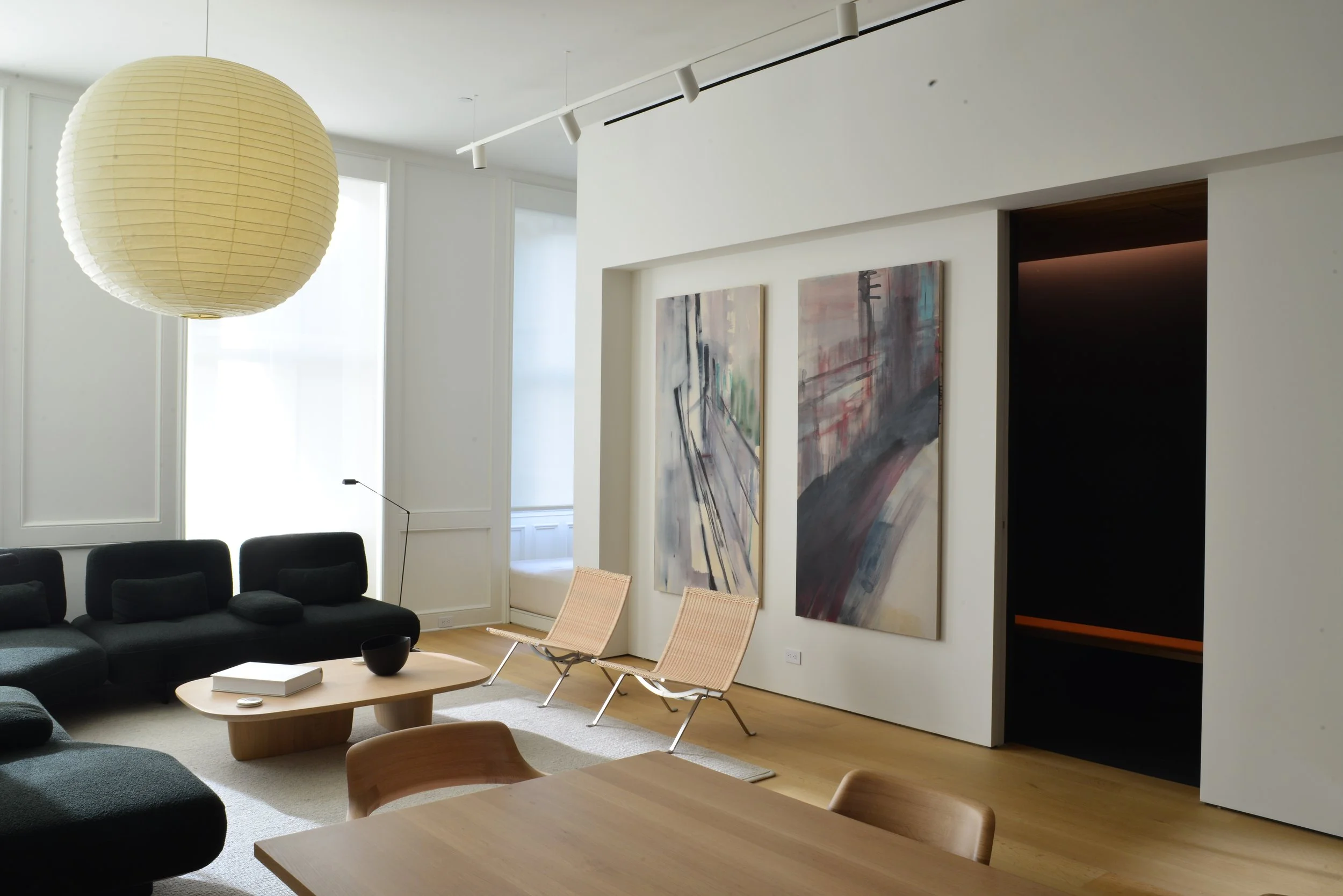 Modern living room with black sofa, light wood coffee table, two beige lounge chairs, abstract paintings, large paper lantern, and natural light coming through the windows.