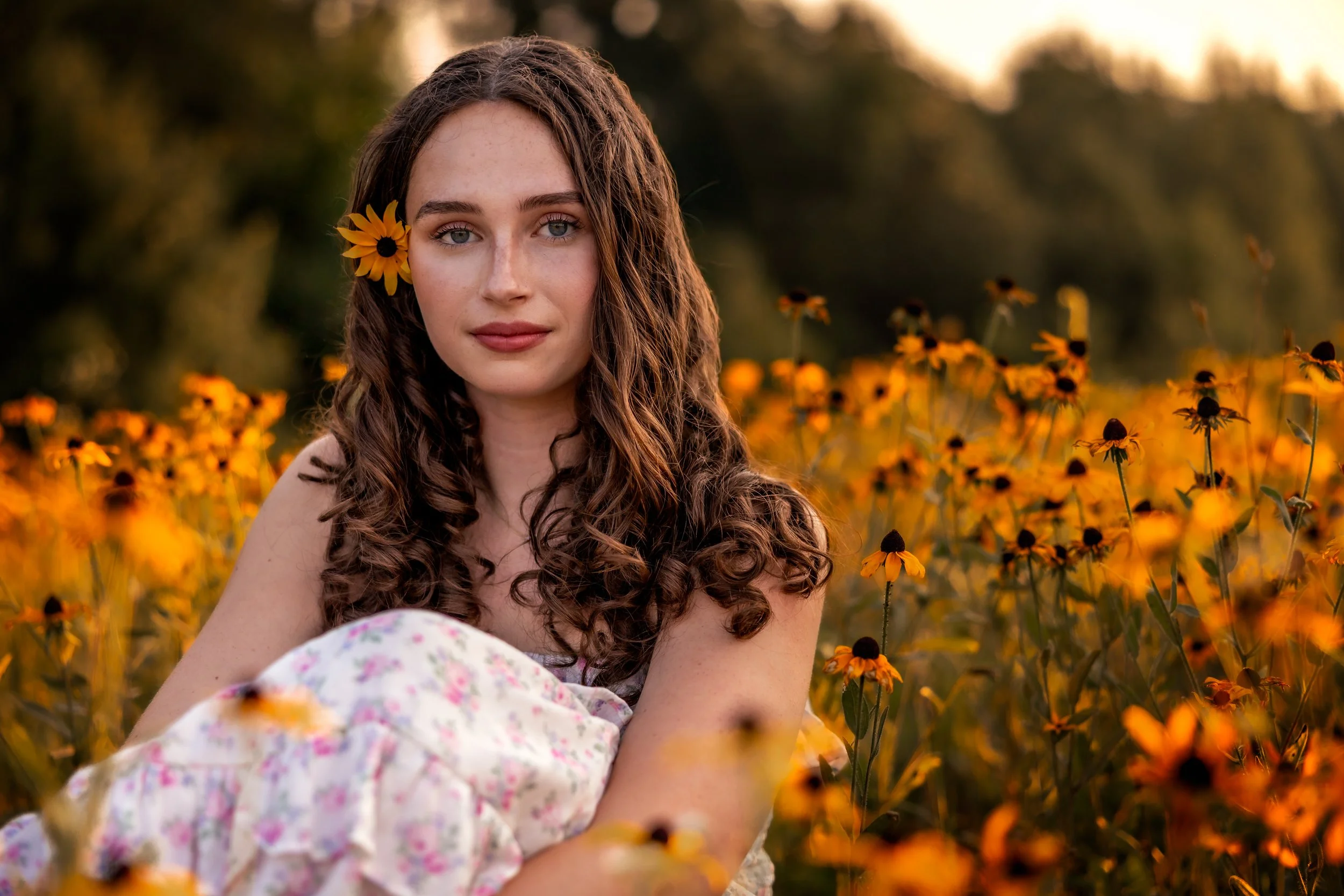 A young woman with long, curly brown hair and blue eyes sitting in a field of yellow flowers with a flower behind her ear, during sunset or golden hour.