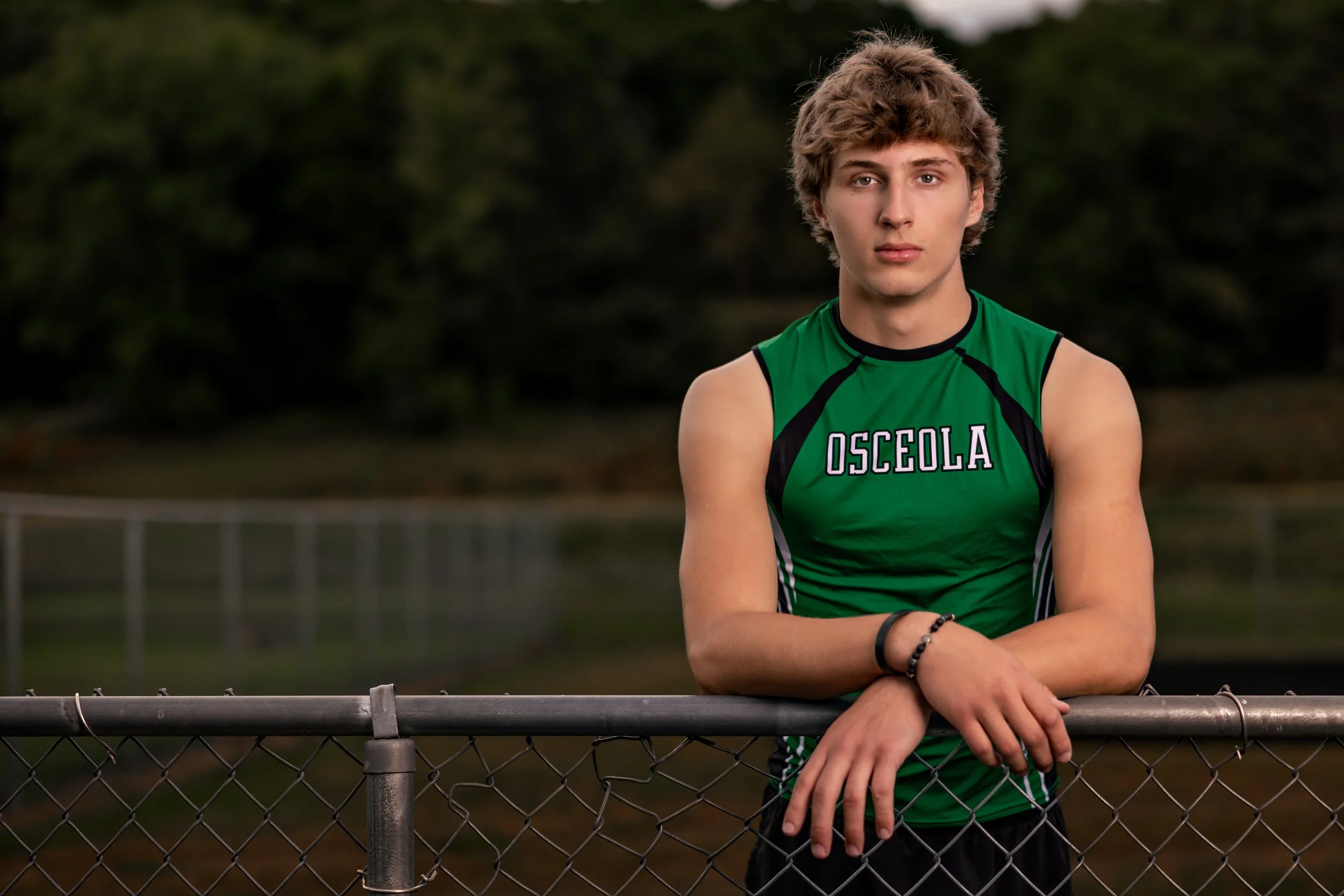 A young man with curly hair wearing a green sports uniform with the word Osceola, standing outdoors behind a chain-link fence on a track or field, with trees in the background.