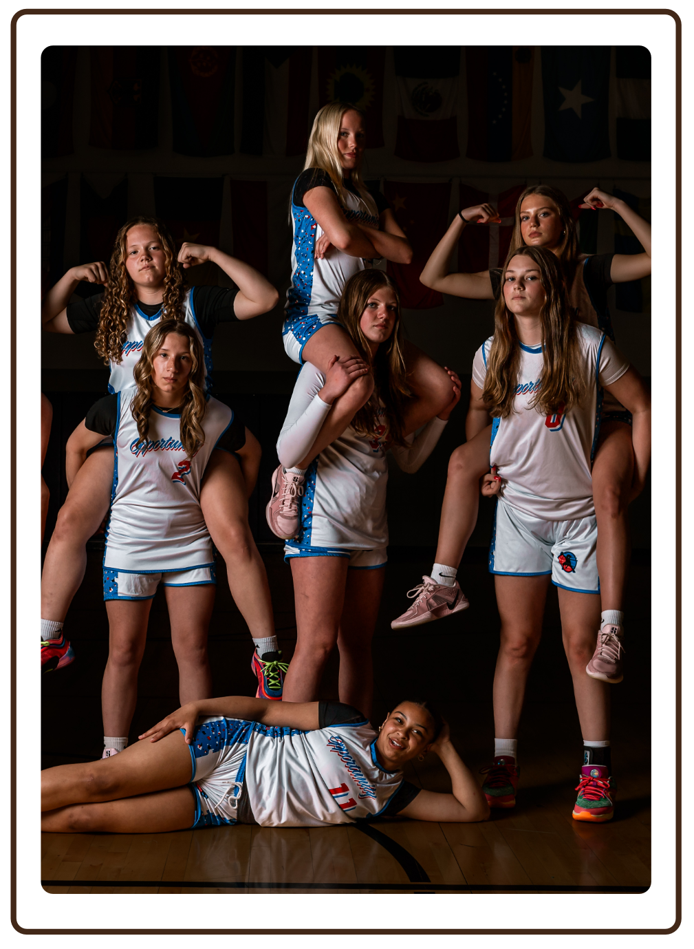 A group of young female basketball players in uniform striking a playful, strong pose, with some flexing muscles and one lying on the floor, in a team photo setting.