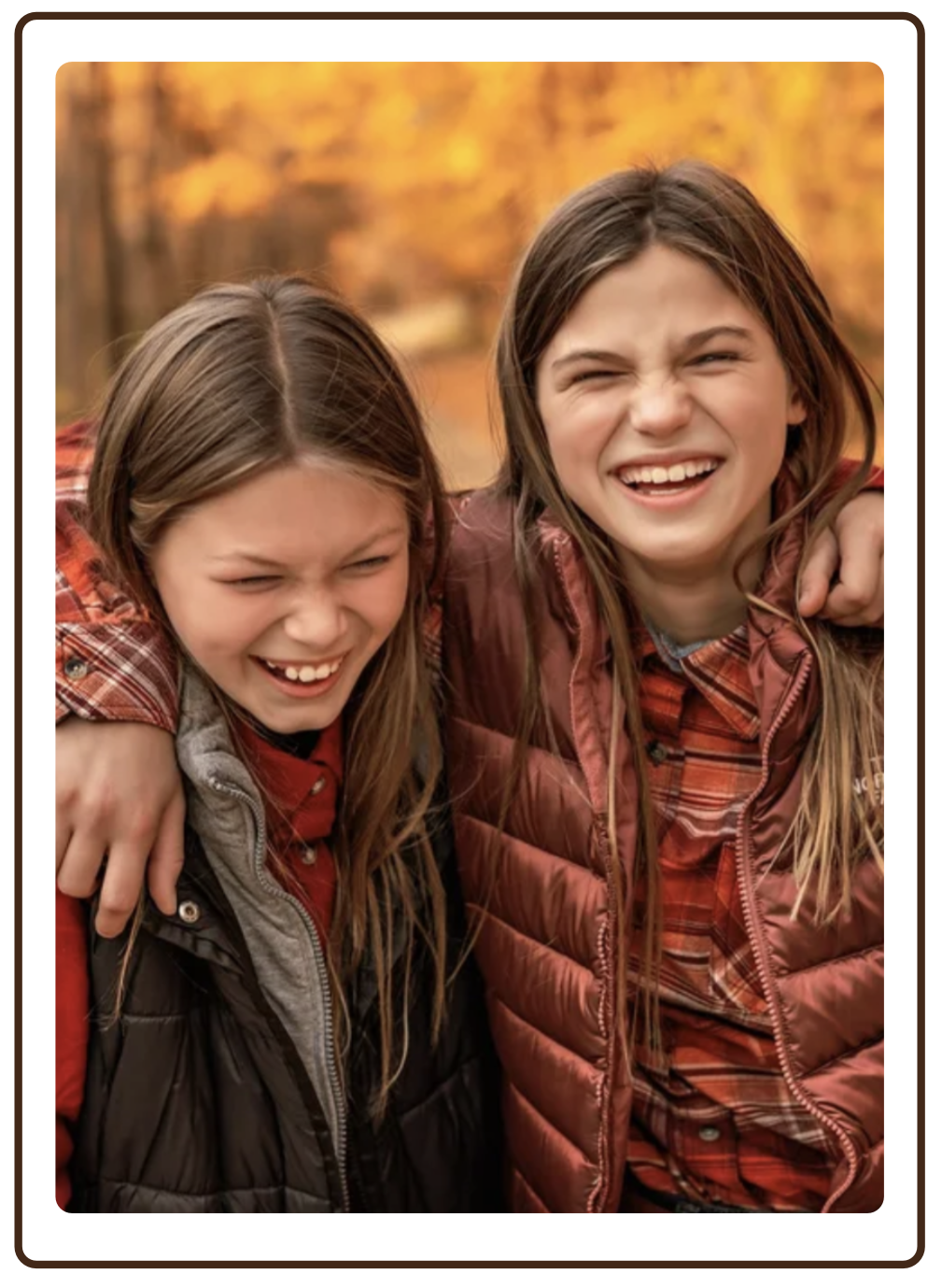 Two young girls smiling and laughing outdoors during autumn, wearing plaid shirts and puffer jackets, with trees in fall foliage in the background.