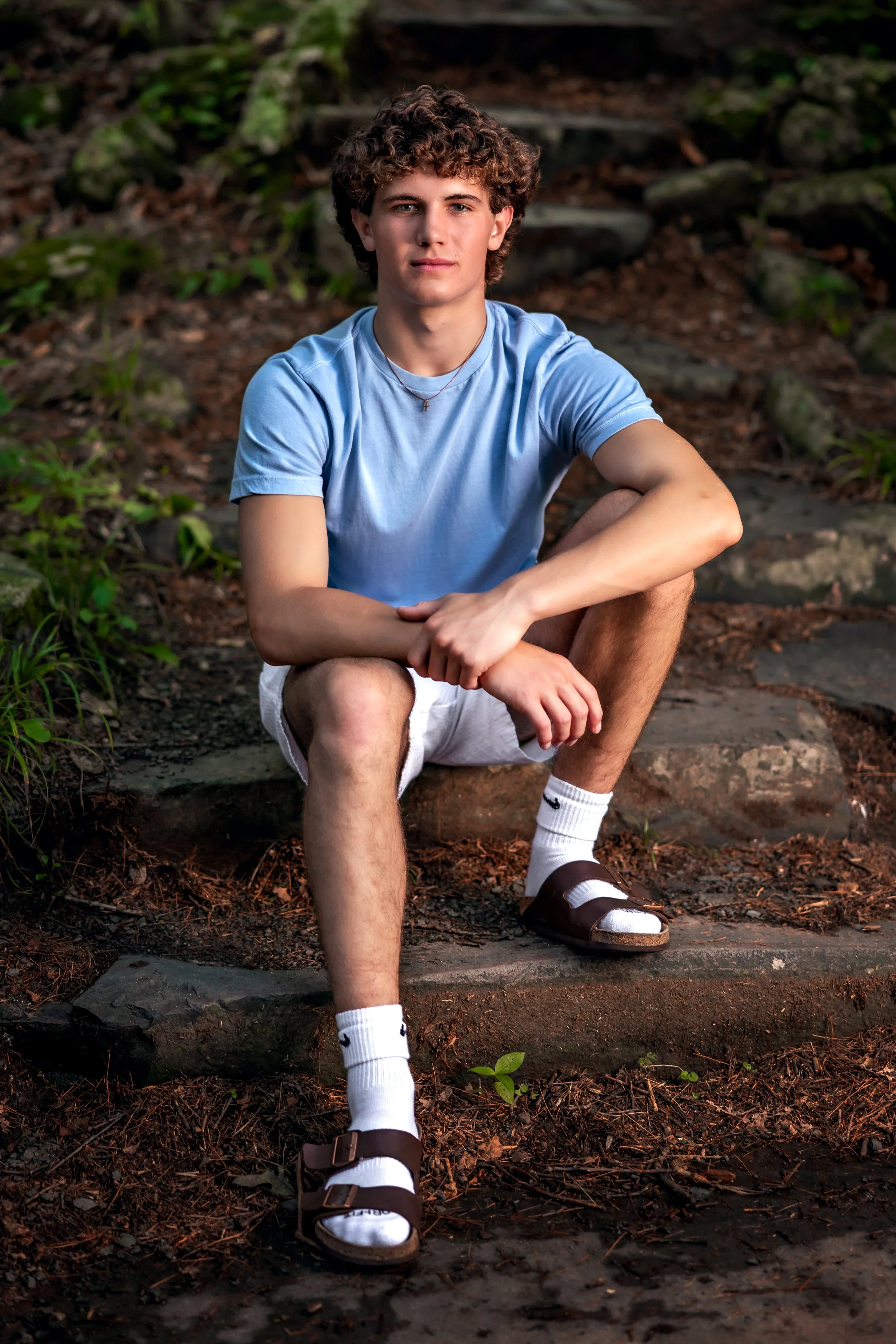 A young man with curly hair sitting outdoors on stone steps surrounded by greenery, wearing a light blue t-shirt, white shorts, white socks, and brown sandals.