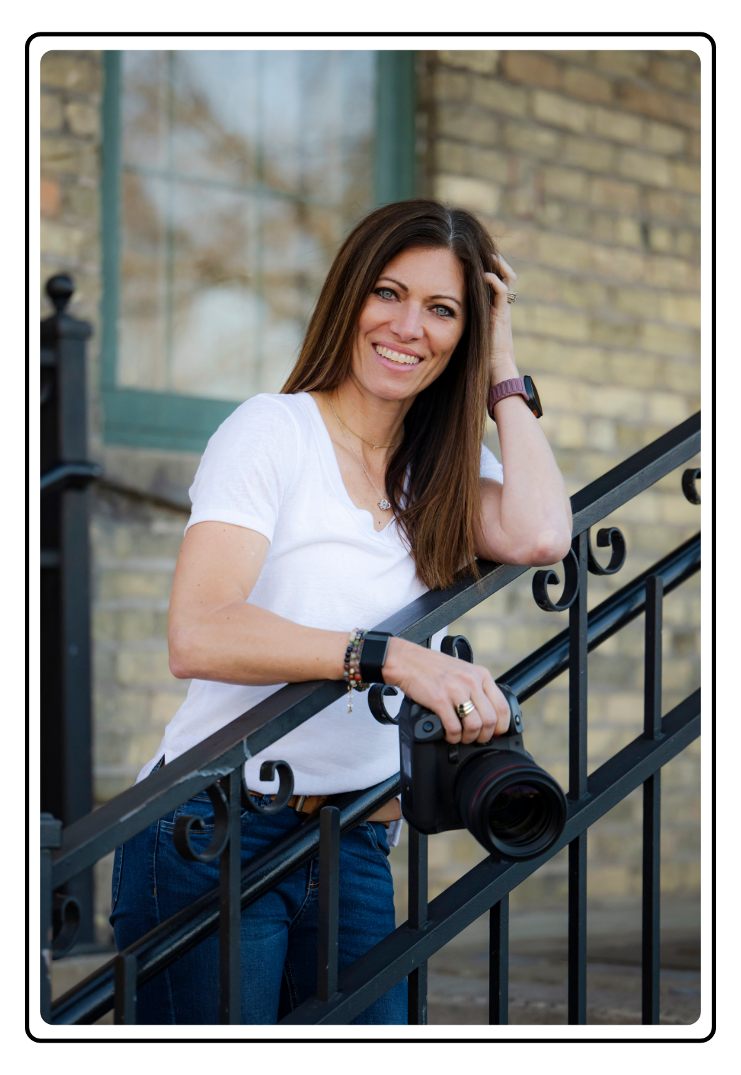 A woman standing on stairs, smiling, with a camera hanging from her hand, wearing a white t-shirt, jeans, and a smartwatch, outdoors with a brick wall and window in the background.