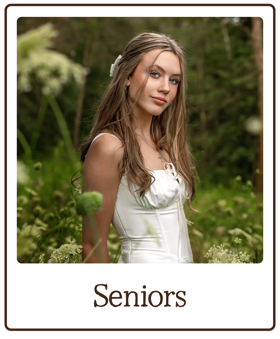A young woman with long wavy hair wearing a white dress, standing outdoors in a green, natural setting, smiling softly at the camera.