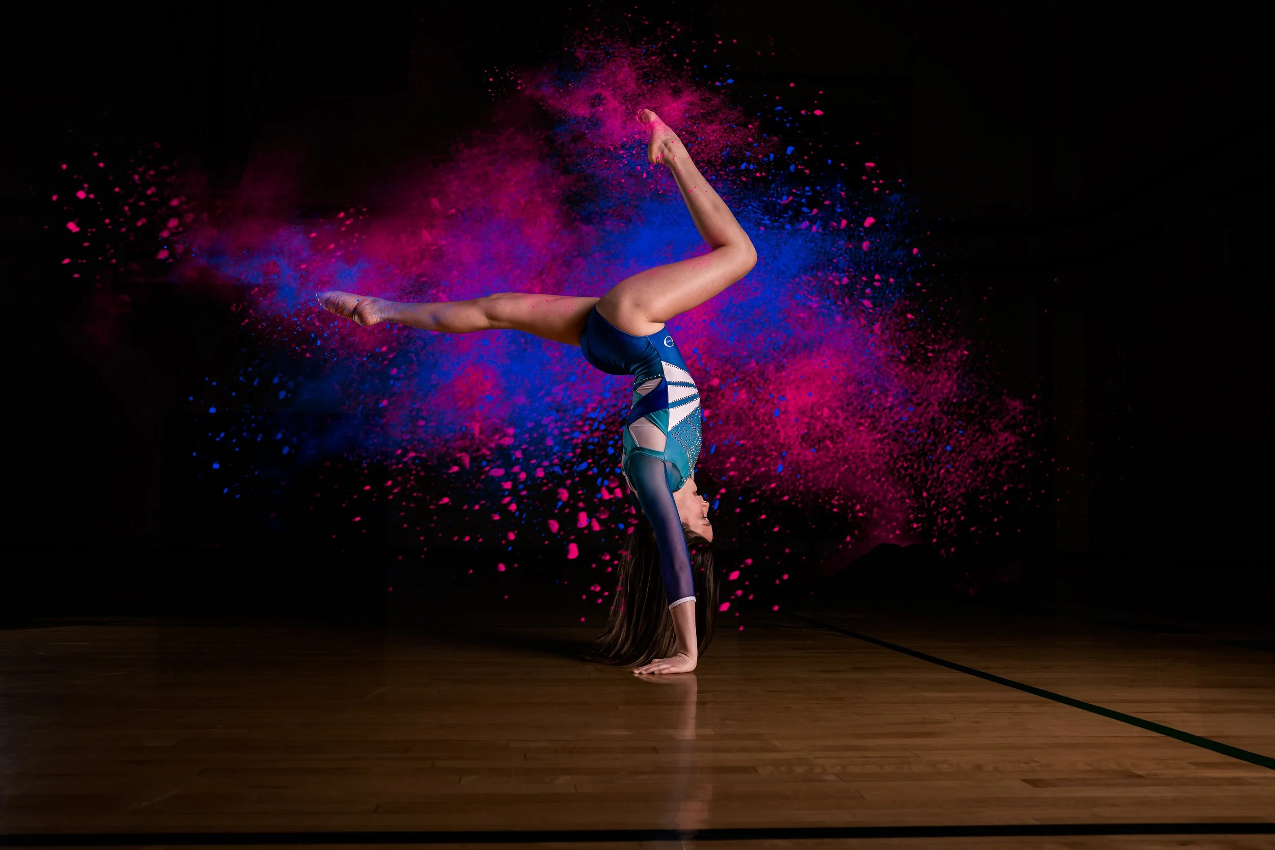 A gymnast performing a handstand on a wooden floor with pink and blue powder exploding behind her against a black background.