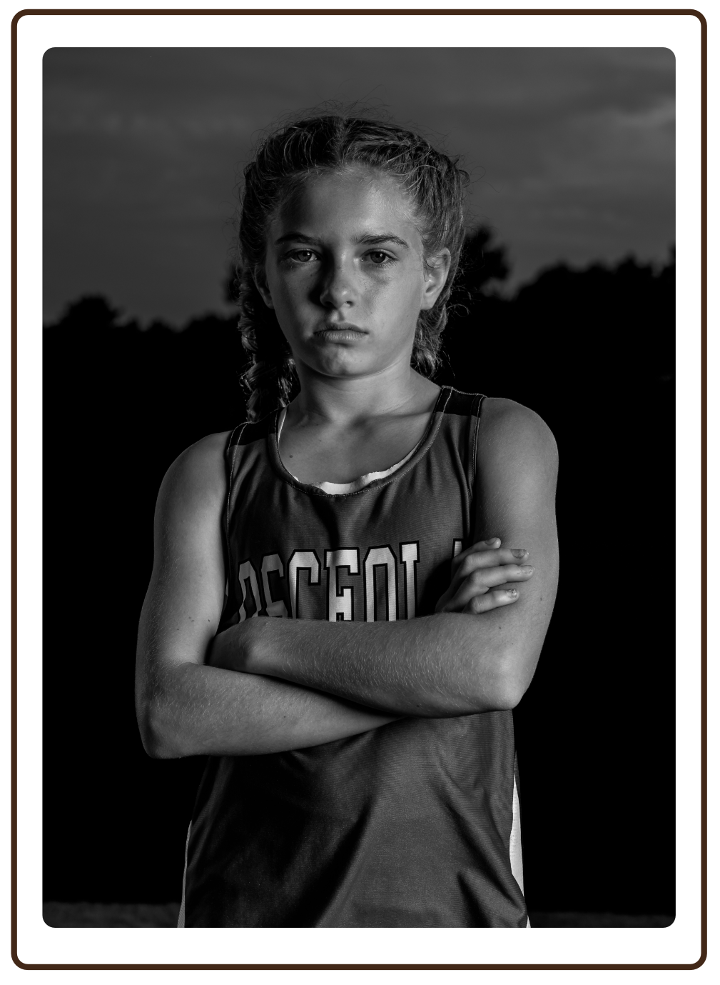 Black and white portrait of a young female athlete with crossed arms, wearing a sports jersey, outdoors at dusk or dawn.