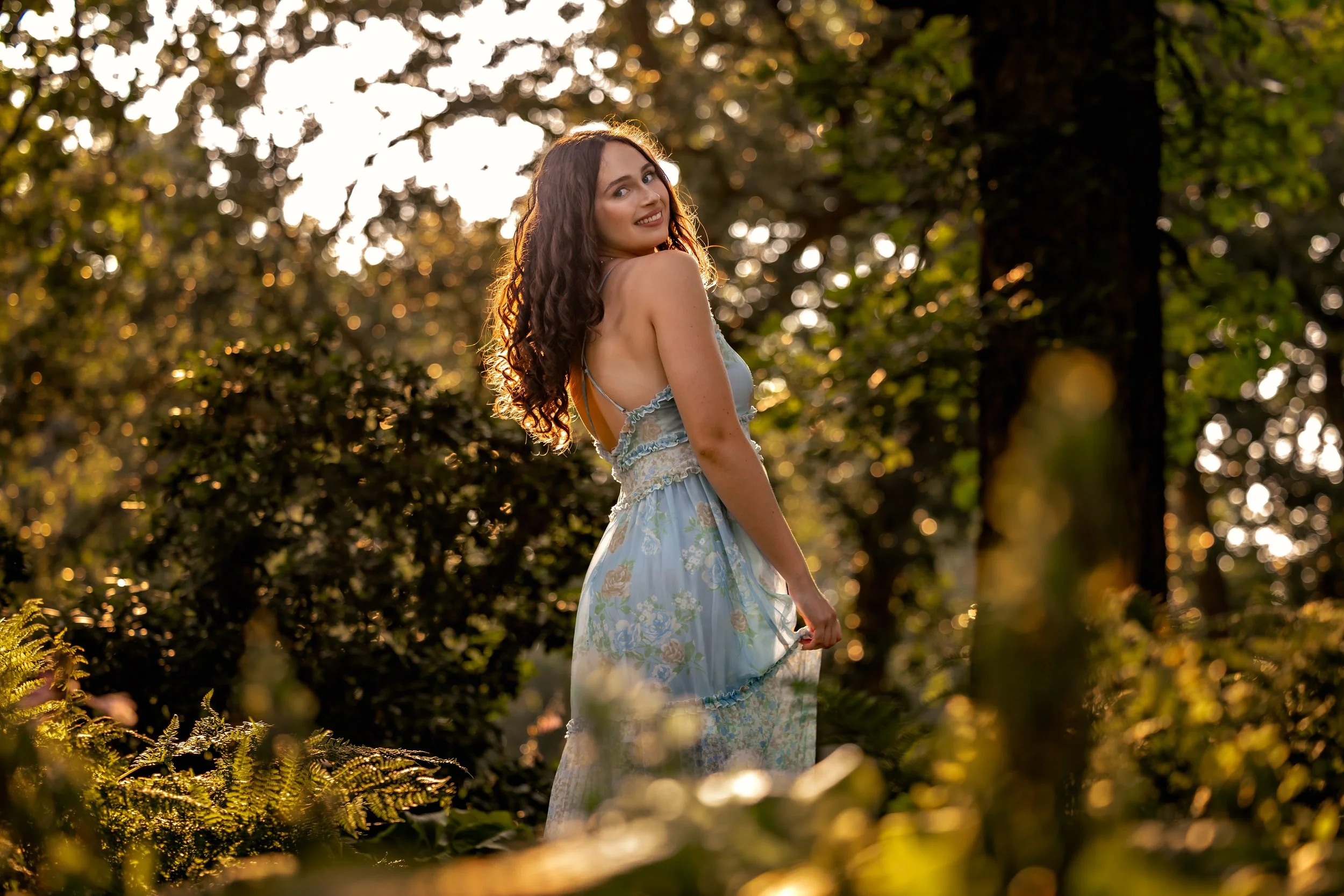 A young woman with long, wavy brown hair wearing a light blue floral dress, standing outdoors in a forest during sunset, smiling and looking back over her shoulder.