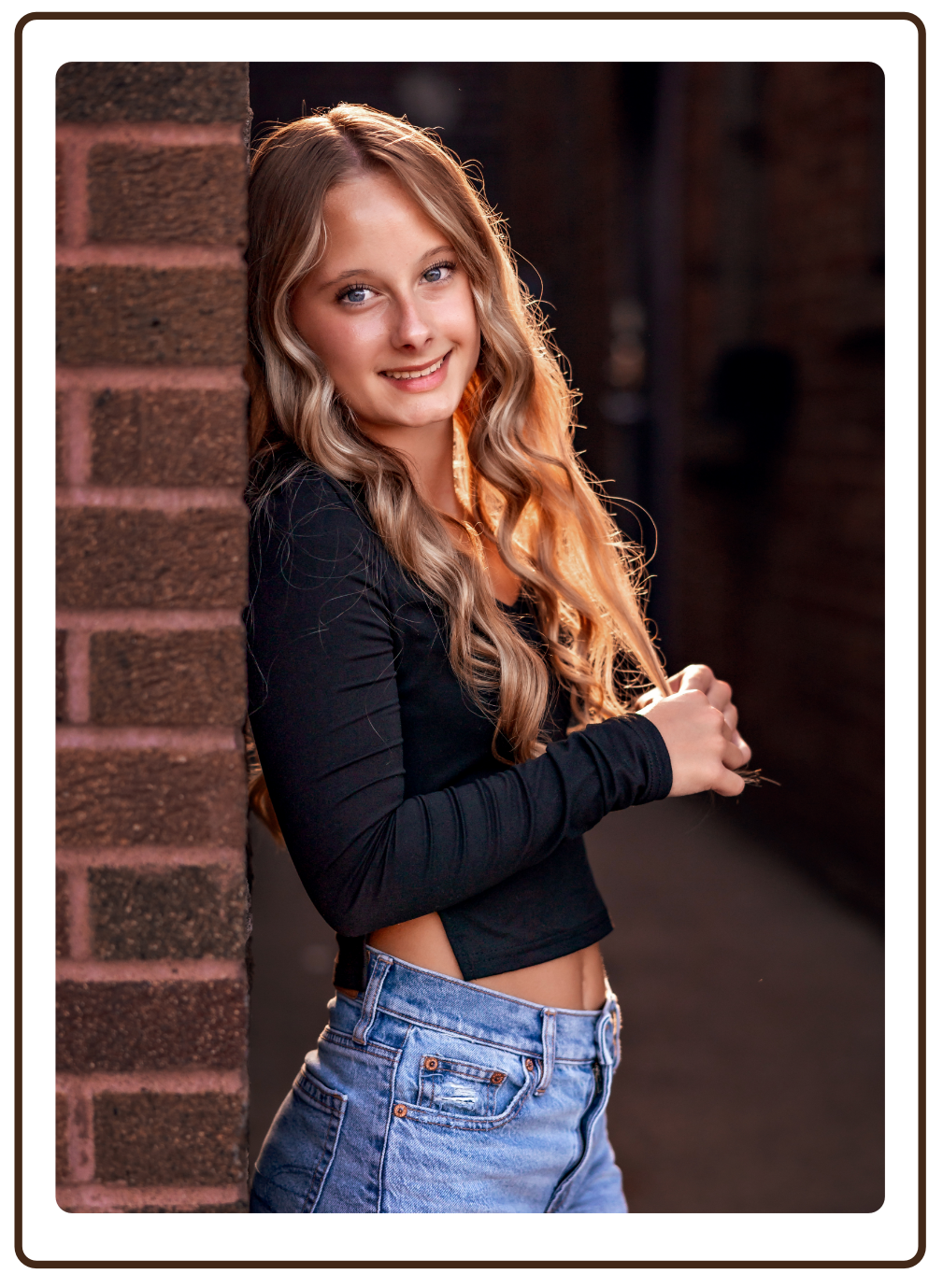 A young woman with long, wavy blond hair, wearing a black long sleeve top and high-waisted blue jeans, smiling and leaning against a brick wall.