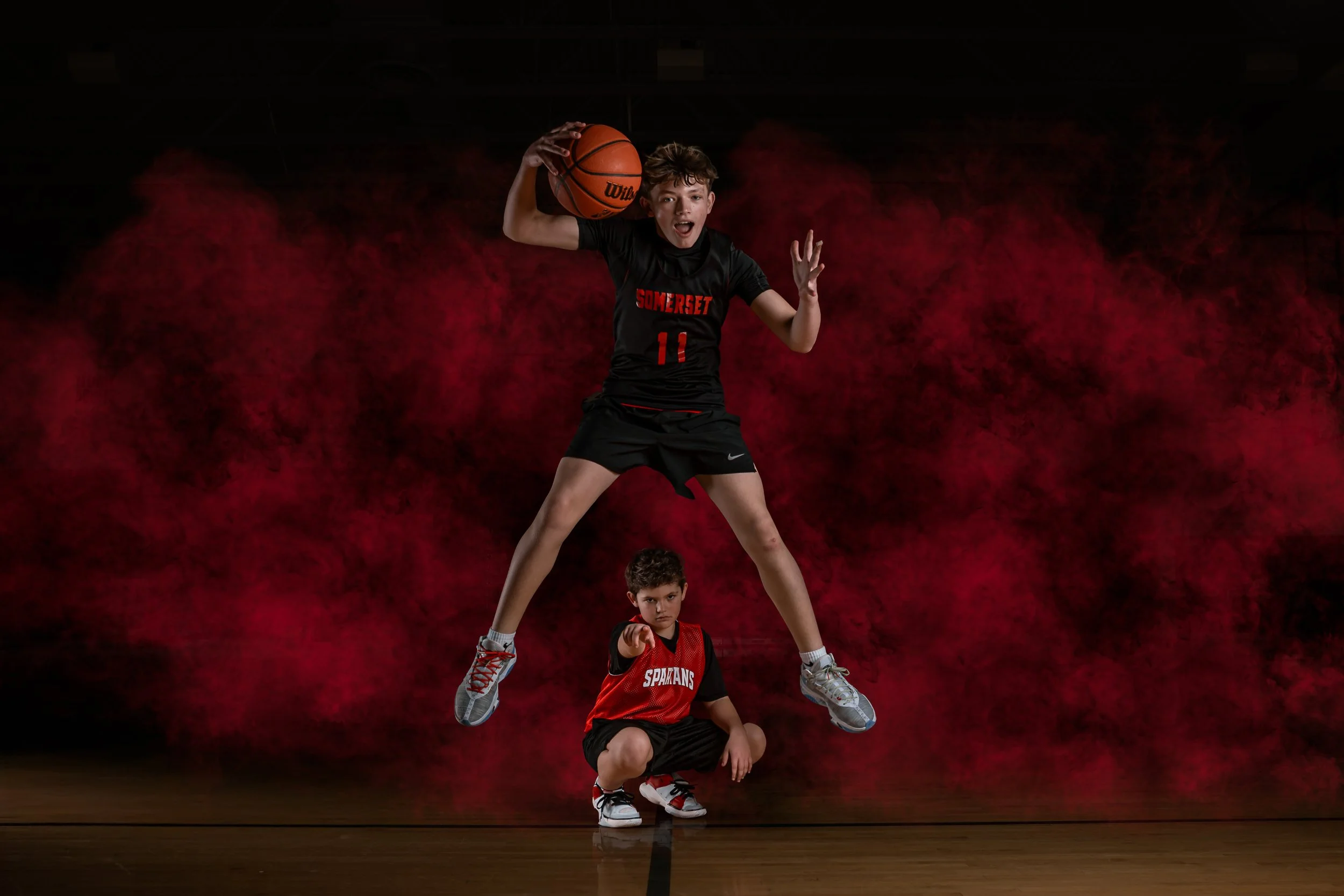 Three boys performing a human pyramid on a basketball court with a dark background and red smoke effects.