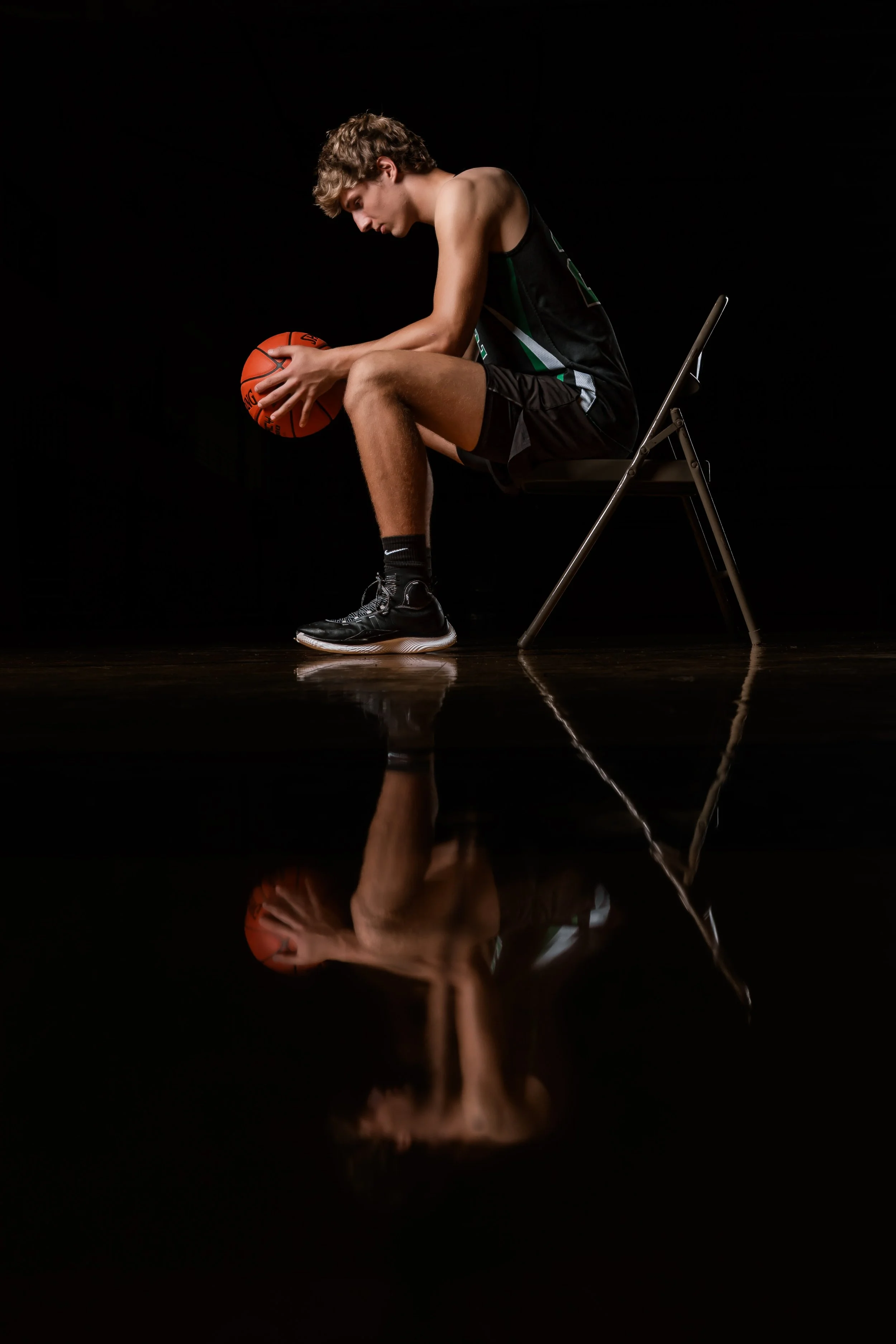 A young male basketball player sitting on a chair in a dark setting, holding a basketball, with his reflection visible on the shiny floor.