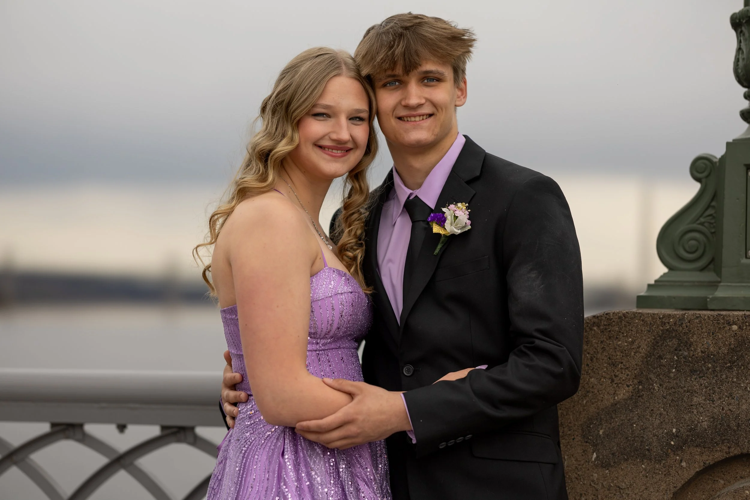 A young couple dressed in formal attire, smiling and embracing each other outdoors during daytime.