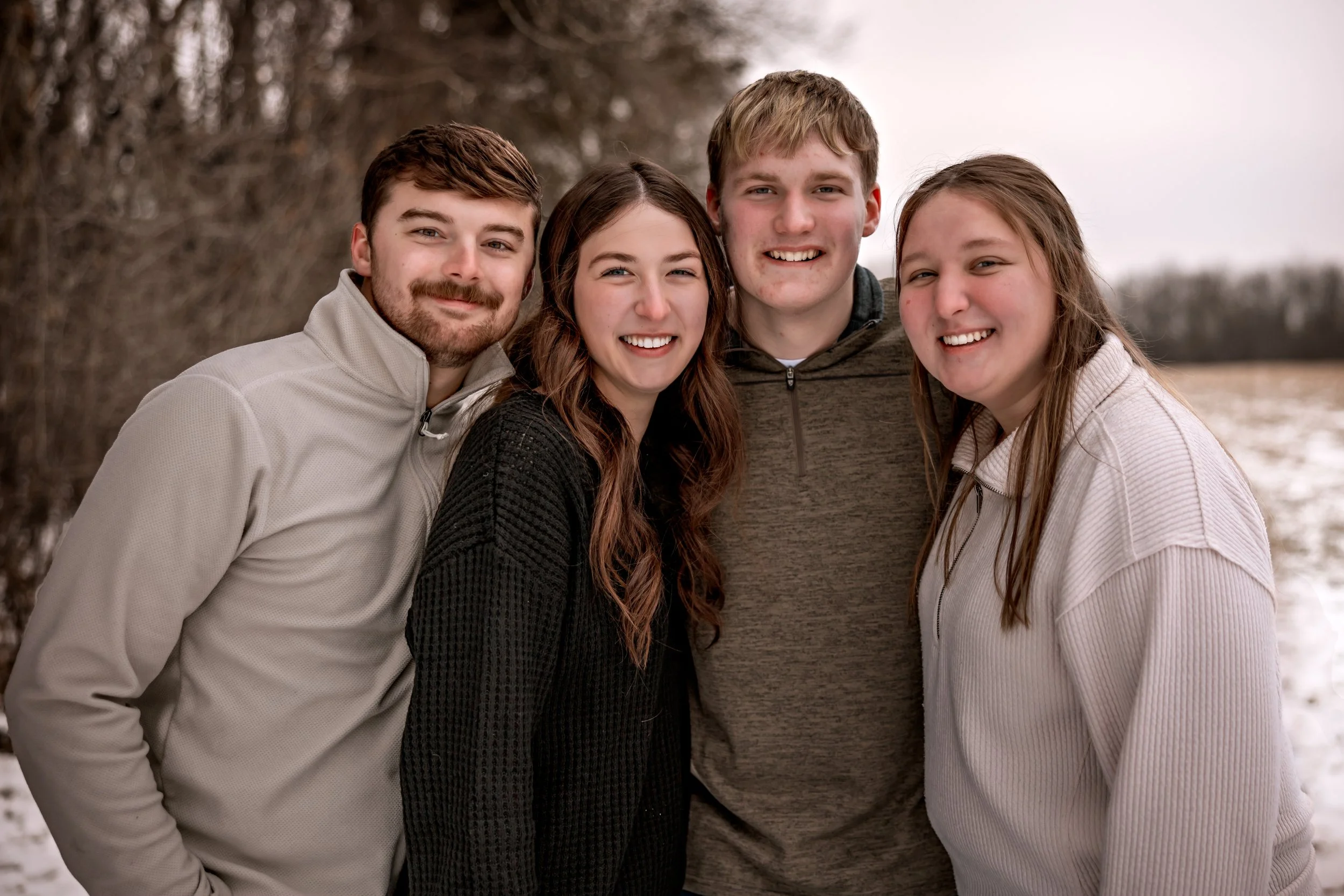 Four young friends smiling outdoors in a winter setting with bare trees and snow, dressed in casual jackets.