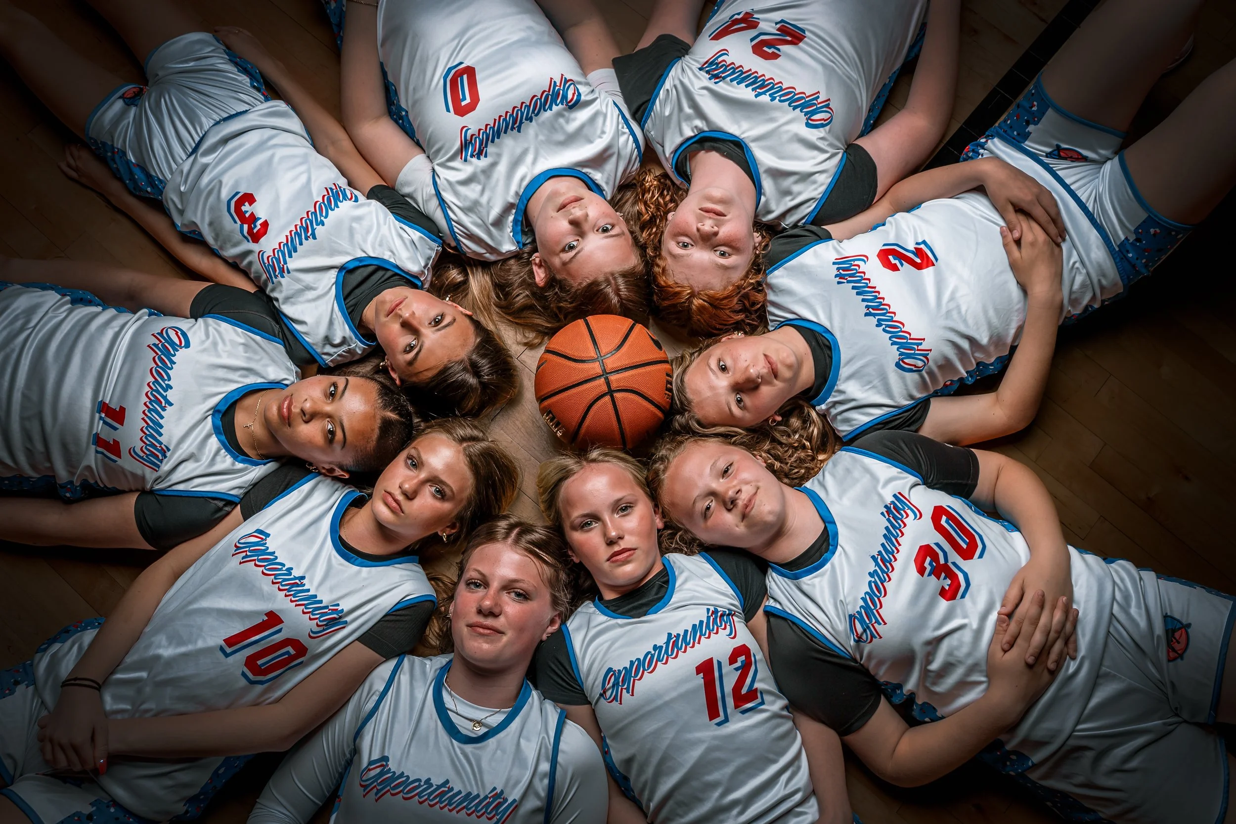 A group of young female basketball players lying on the floor in a circle, wearing white jerseys with blue and red accents, and a basketball in the center.