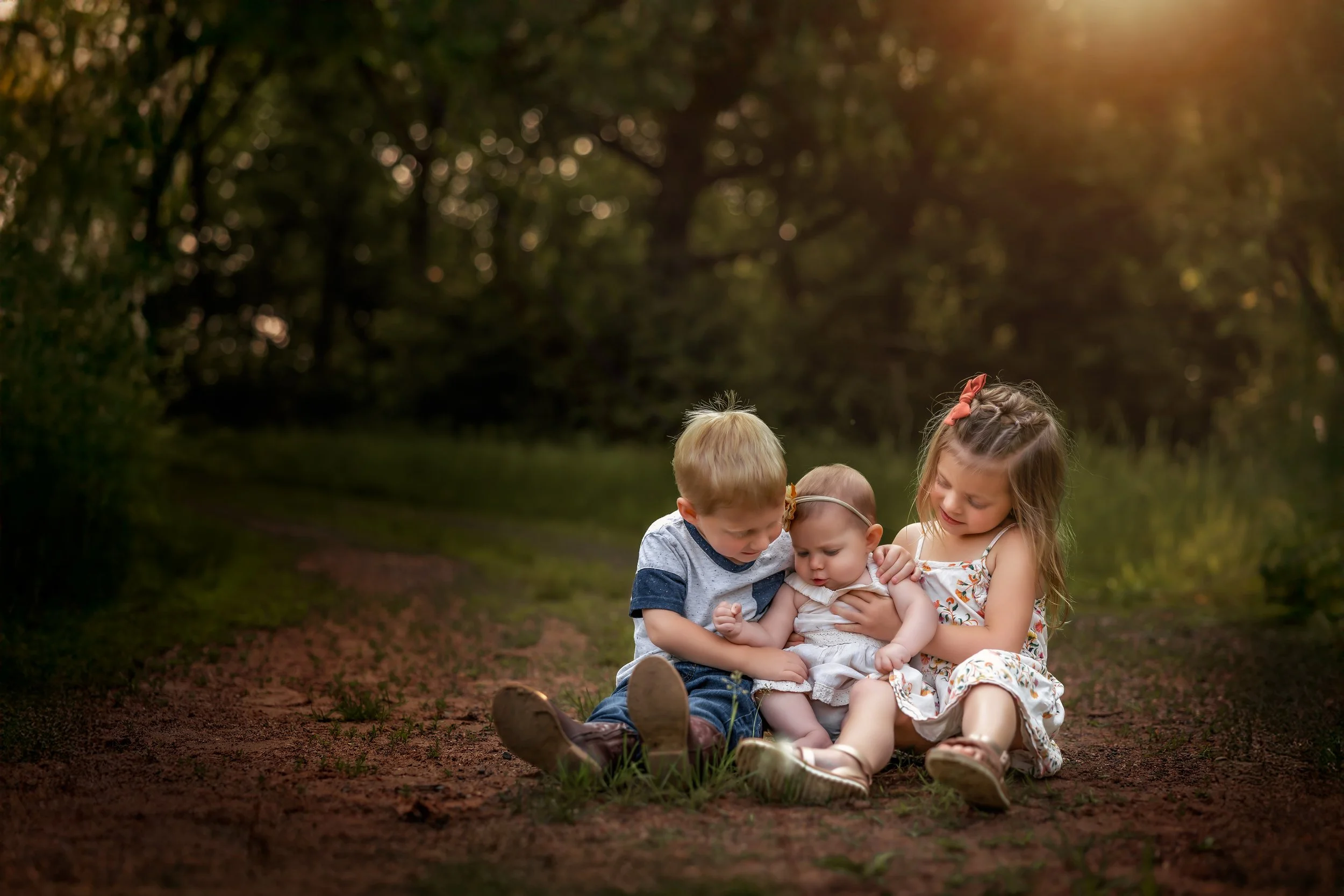 Four children sit together on a dirt path outdoors during sunset, with trees in the background, examining something in their hands.
