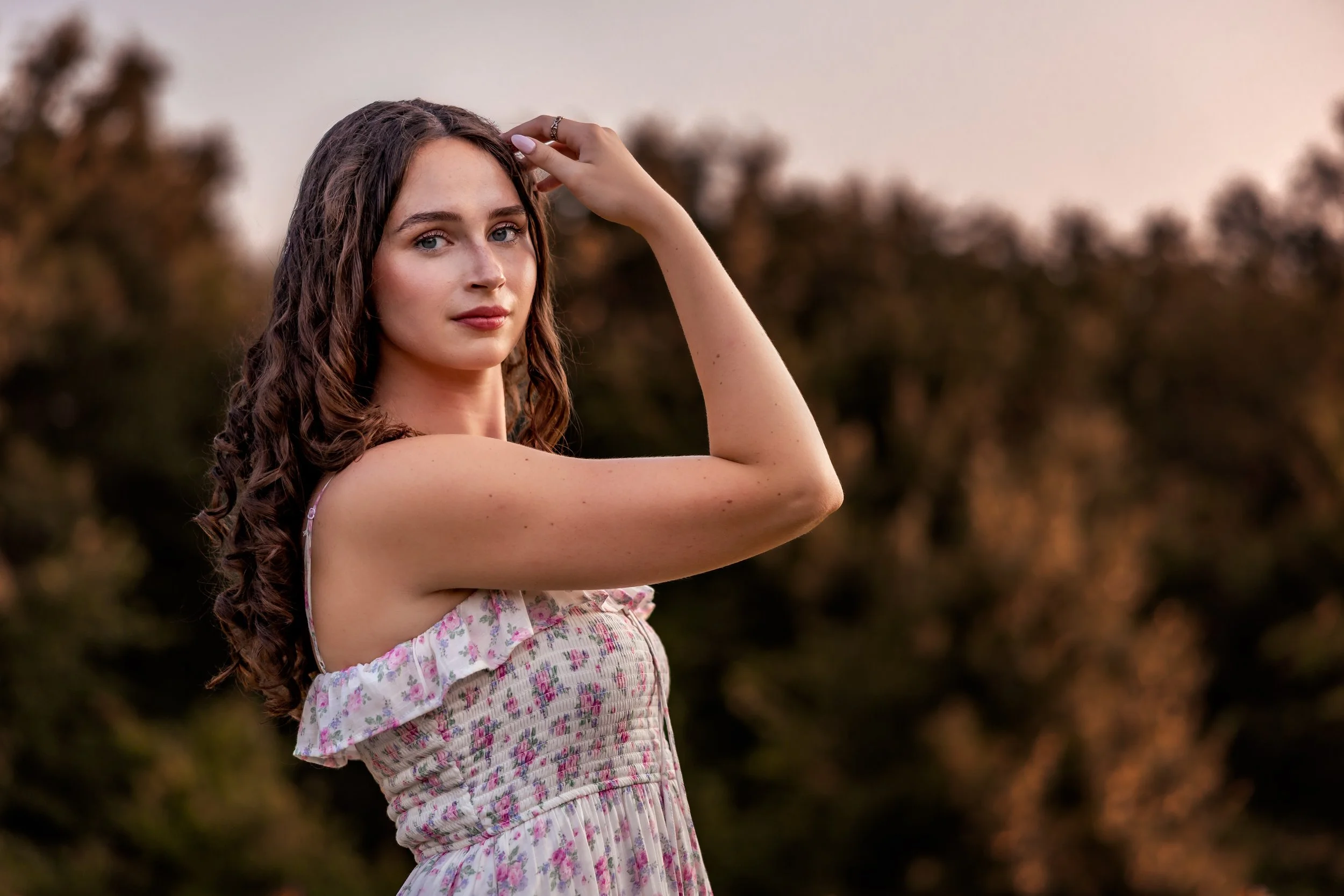 A young woman with long, curly brown hair and blue eyes is standing outdoors during sunset, wearing a floral, off-shoulder dress, with her right arm raised and hand resting on her head, looking at the camera with a slight smile.