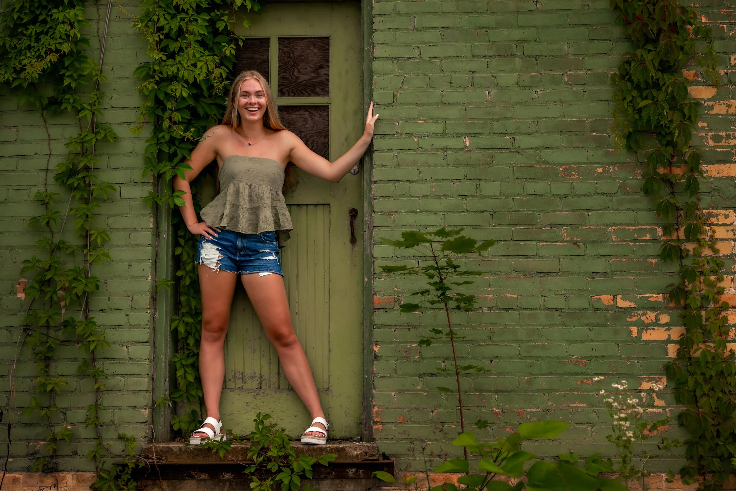 A young woman with long blonde hair, wearing a strapless beige top, ripped denim shorts, and white sandals, is smiling and standing in front of a green brick wall partially covered with green vines. She is leaning against a green door with windows, h