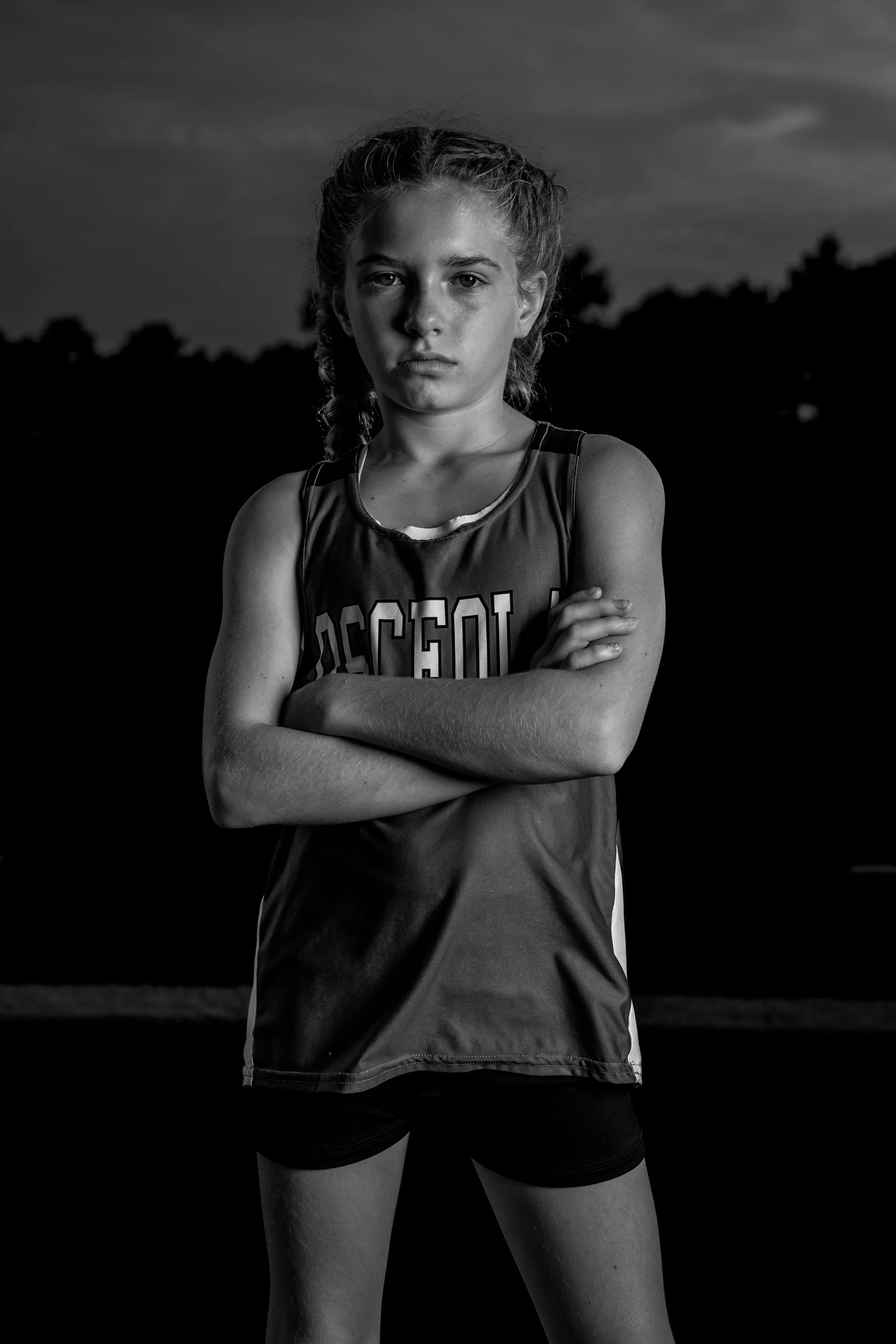 Black and white photo of a young female athlete standing outdoors with her arms crossed, wearing sportswear that reads 'CrossFit', with trees and a cloudy sky in the background.