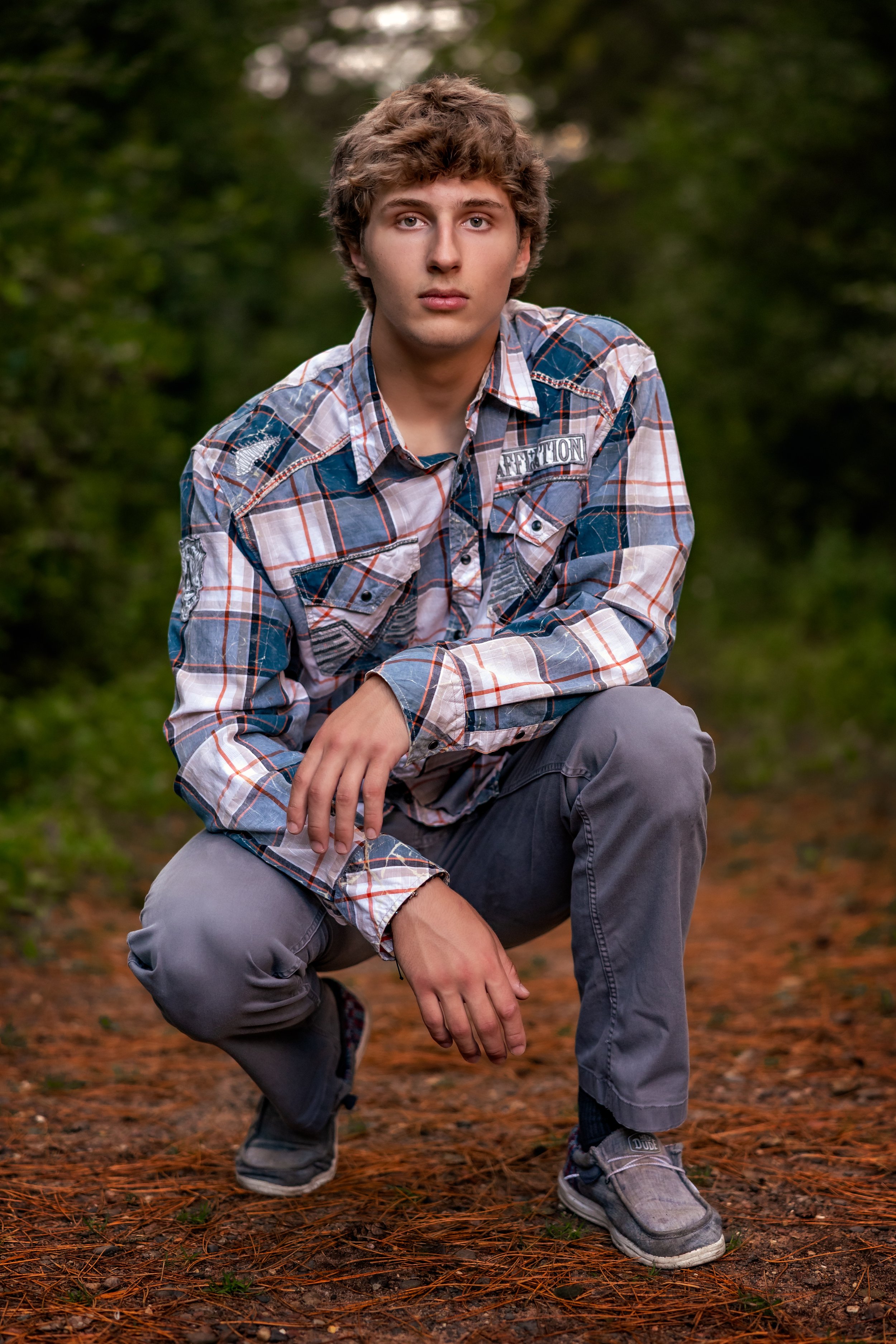 A young man is kneeling outdoors on a forest trail with trees and pine needles in the background, wearing a plaid shirt, gray pants and sneakers.