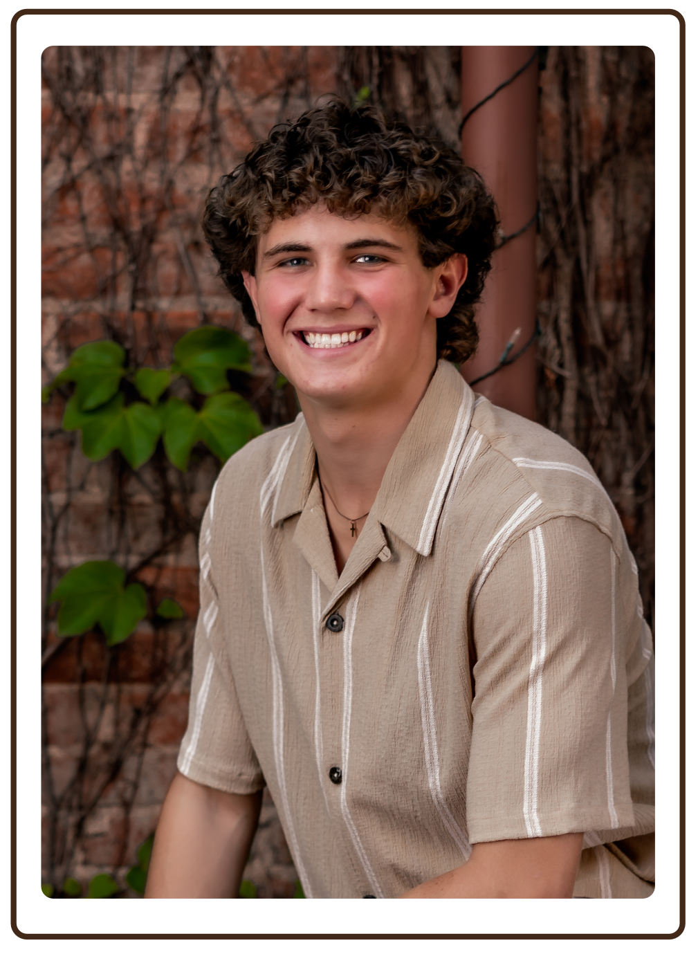 A young man with curly hair smiling, wearing a beige striped short-sleeve shirt, standing outdoors in front of a tree with ivy and vines.