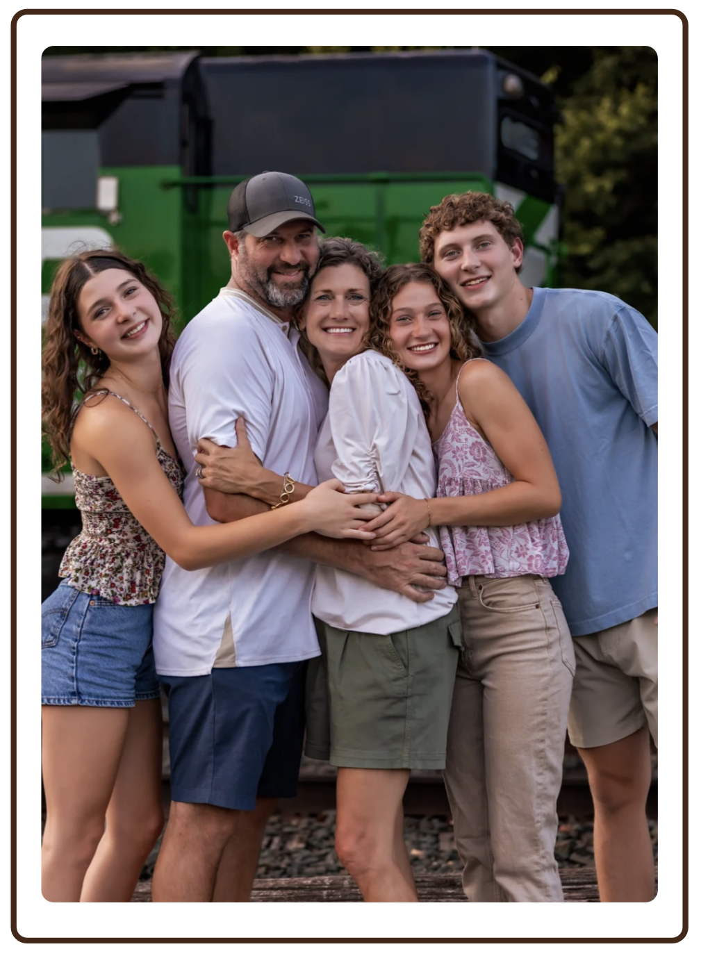 A family of five, including two young women, an older woman, a man, and a teenage boy, embracing and smiling outdoors with a green locomotive in the background.