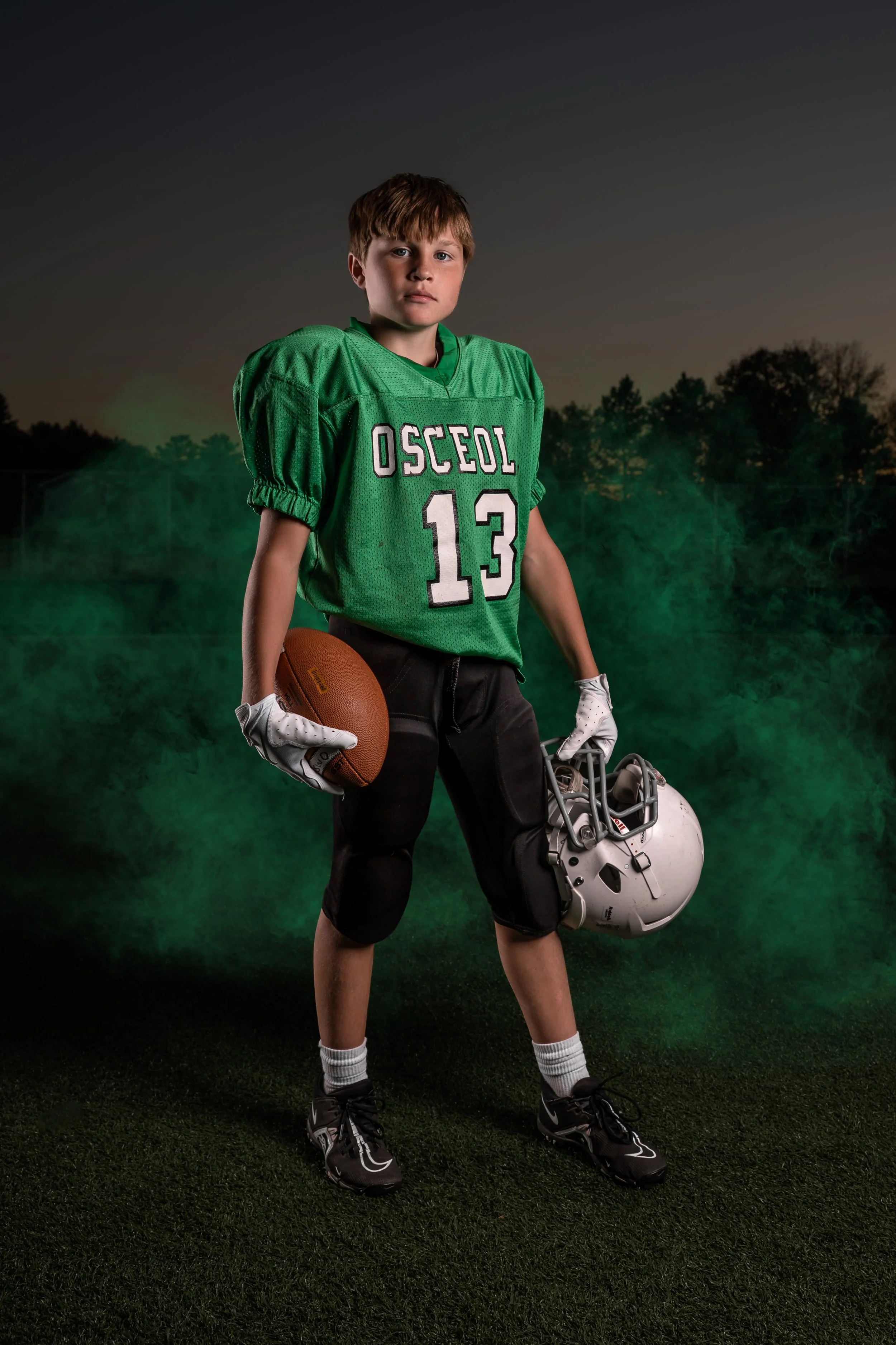 Young football player in green Osceol uniform holding a football and helmet on a football field during evening with a dark sky and trees in the background.