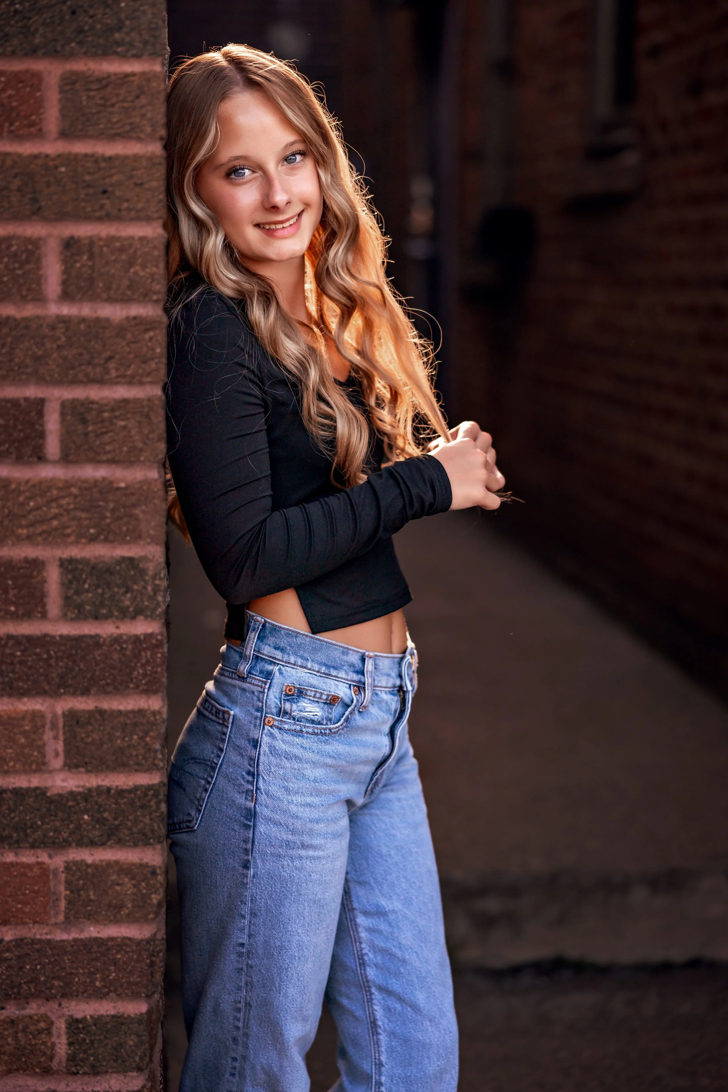 A young woman with long, wavy blonde hair and blue eyes smiling while leaning against a brick wall in an alleyway, wearing a black long-sleeve crop top and blue jeans.