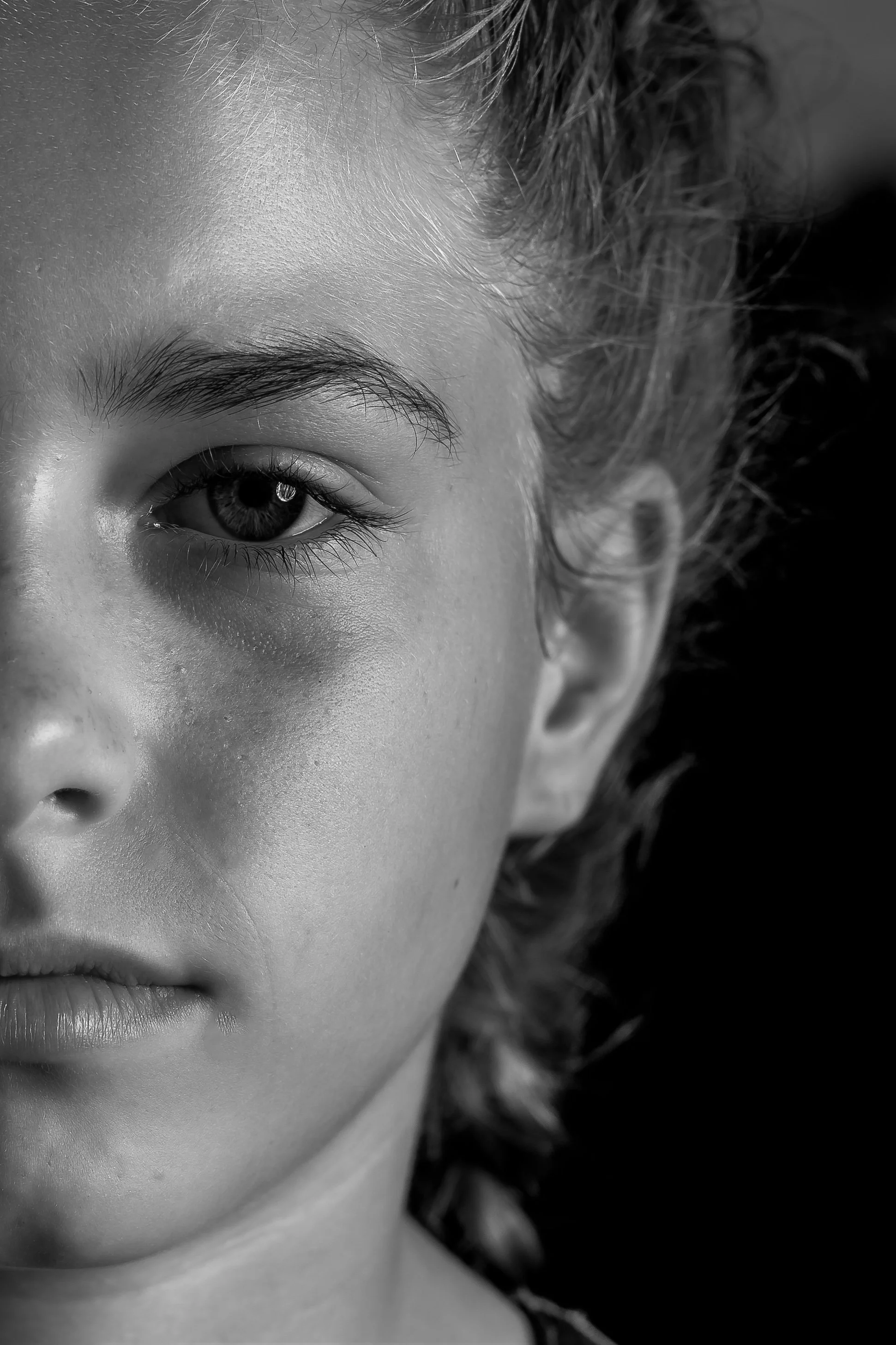 Close-up black and white photo of a person's face, showing an eye, eyebrow, part of the nose, and some facial skin and hair.