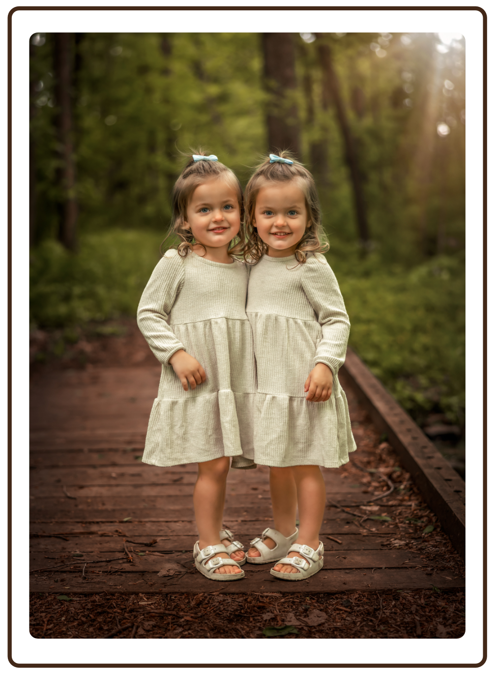 Two young girls with curly hair and bow hair accessories standing close together on a wooden bridge in a forested area, smiling at the camera.