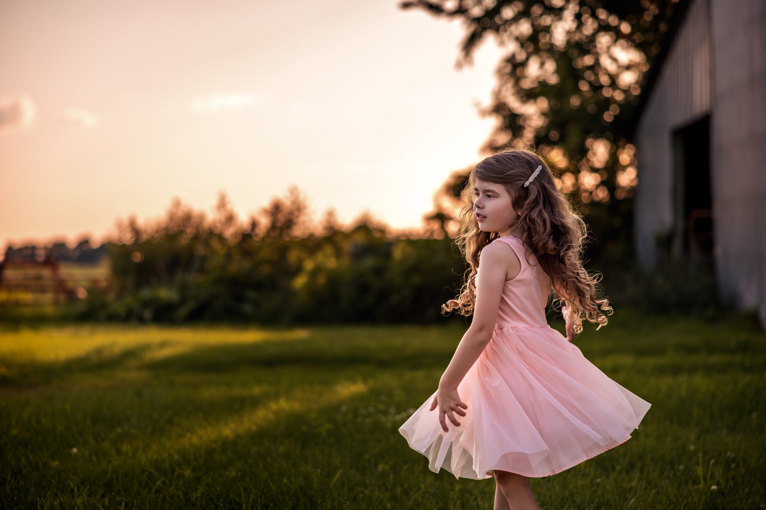 A young girl with long, curly brown hair and a white hair clip, wearing a pink dress, spinning in a grassy outdoor area at sunset.