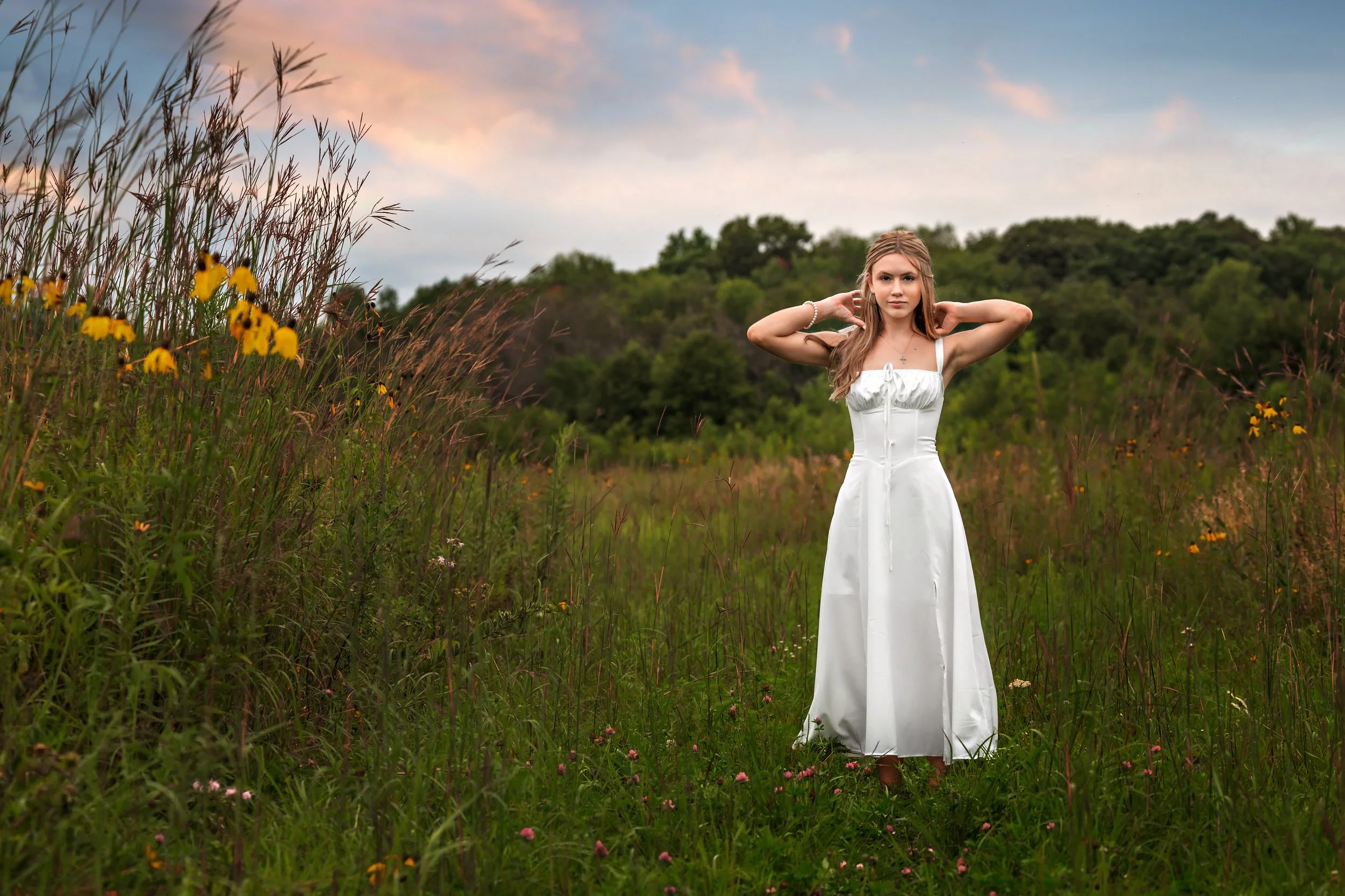 Young woman in a white dress standing in a grassy field with wildflowers, with trees in the background and a cloudy sky.
