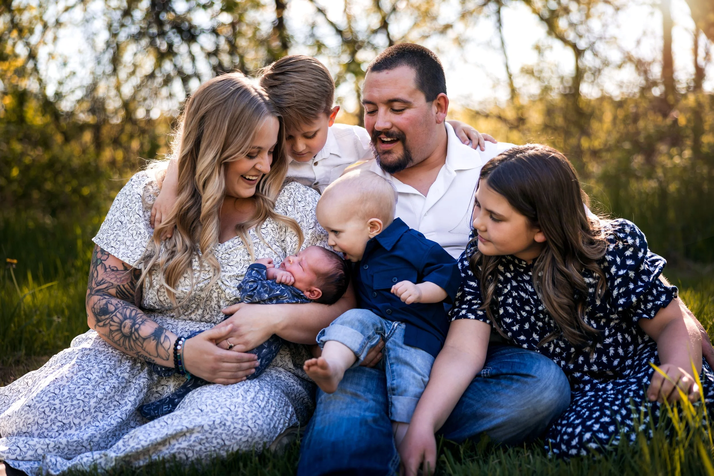 A family of six, including a woman with tattoos, a man, two girls, a boy, and a baby, sitting on grass outdoors during sunset, sharing a joyful moment.