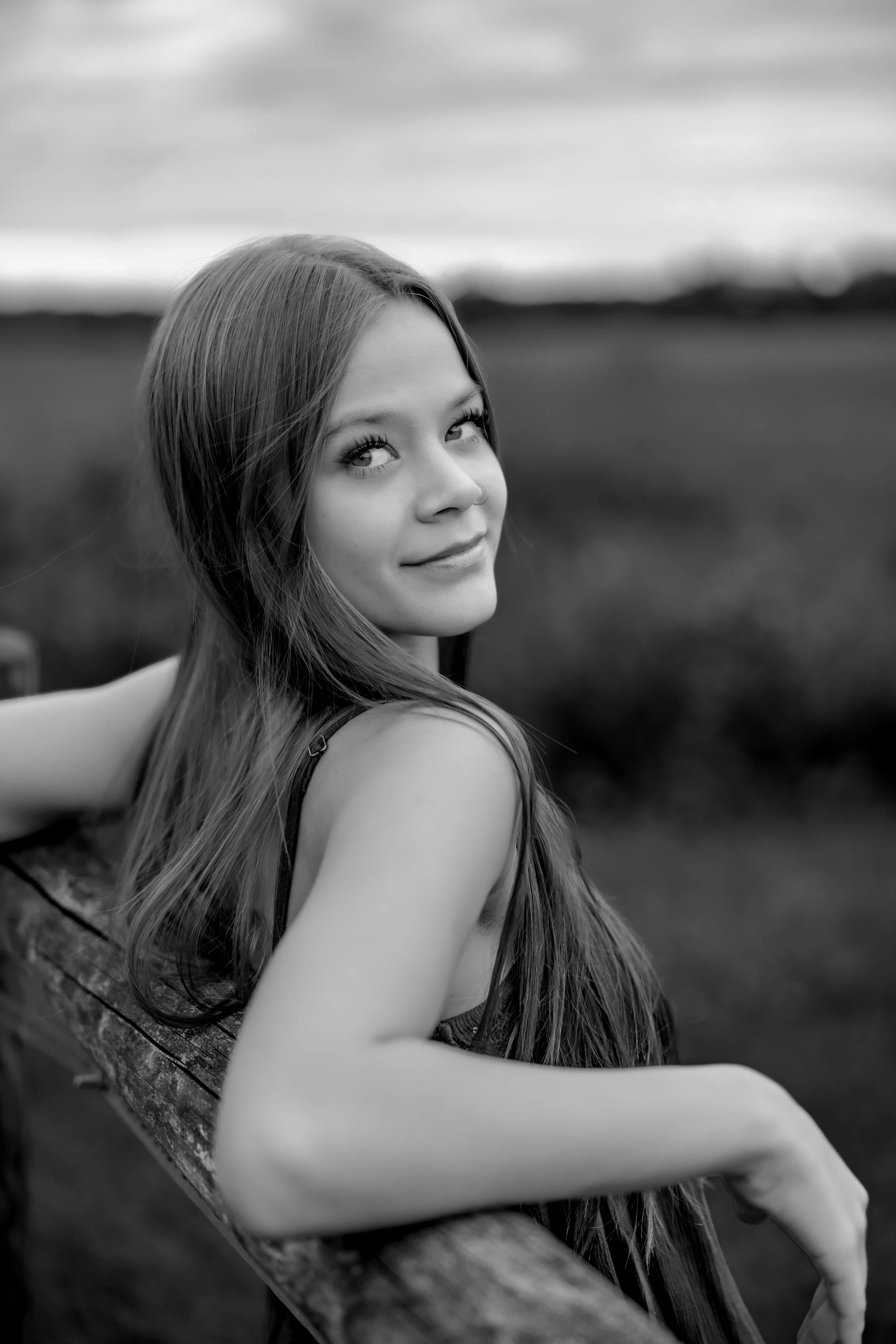 Black and white photo of a young woman with long hair smiling outdoors, sitting on a wooden bench, with a blurred natural background.