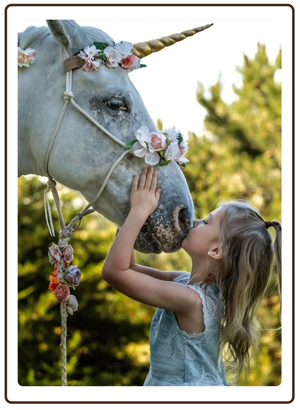 A young girl is giving a kiss and hug to a white unicorn with a golden horn, decorated with pink and white flowers, outdoors with trees in the background.