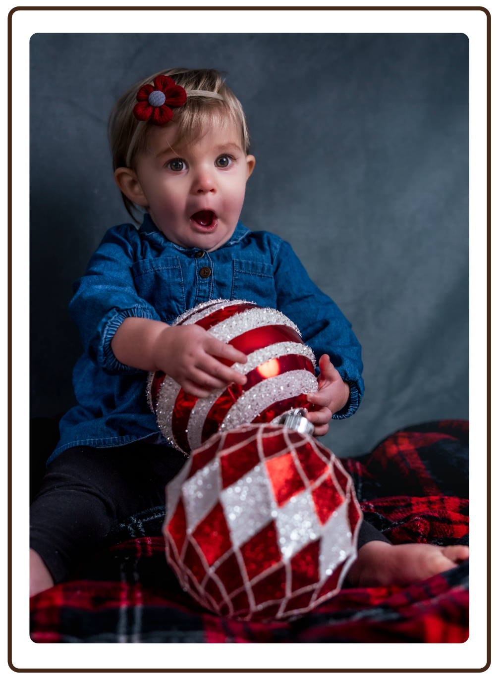 A young girl with a surprised expression, wearing a blue denim shirt and a red flower hair clip, holding a large Christmas ornament, sitting on a red and black plaid blanket.