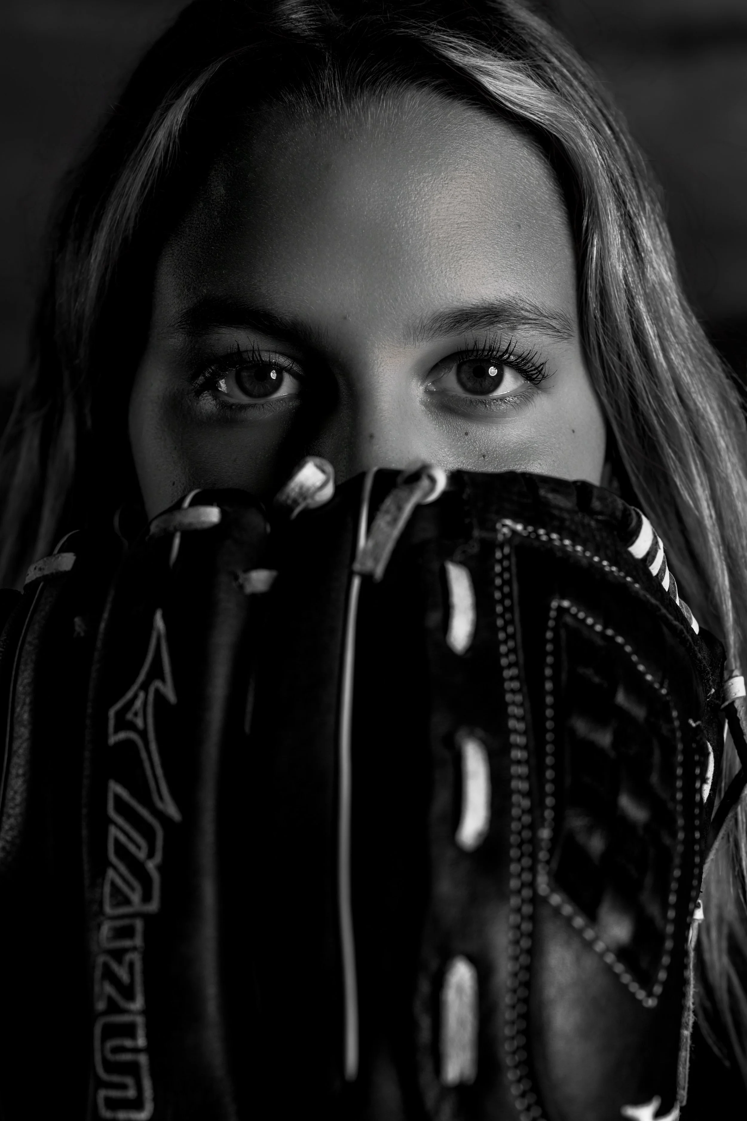 Close-up black and white photo of a young woman's face, with focus on her blue eyes, smooth forehead, and partly covered face by her hand and a jacket with text.