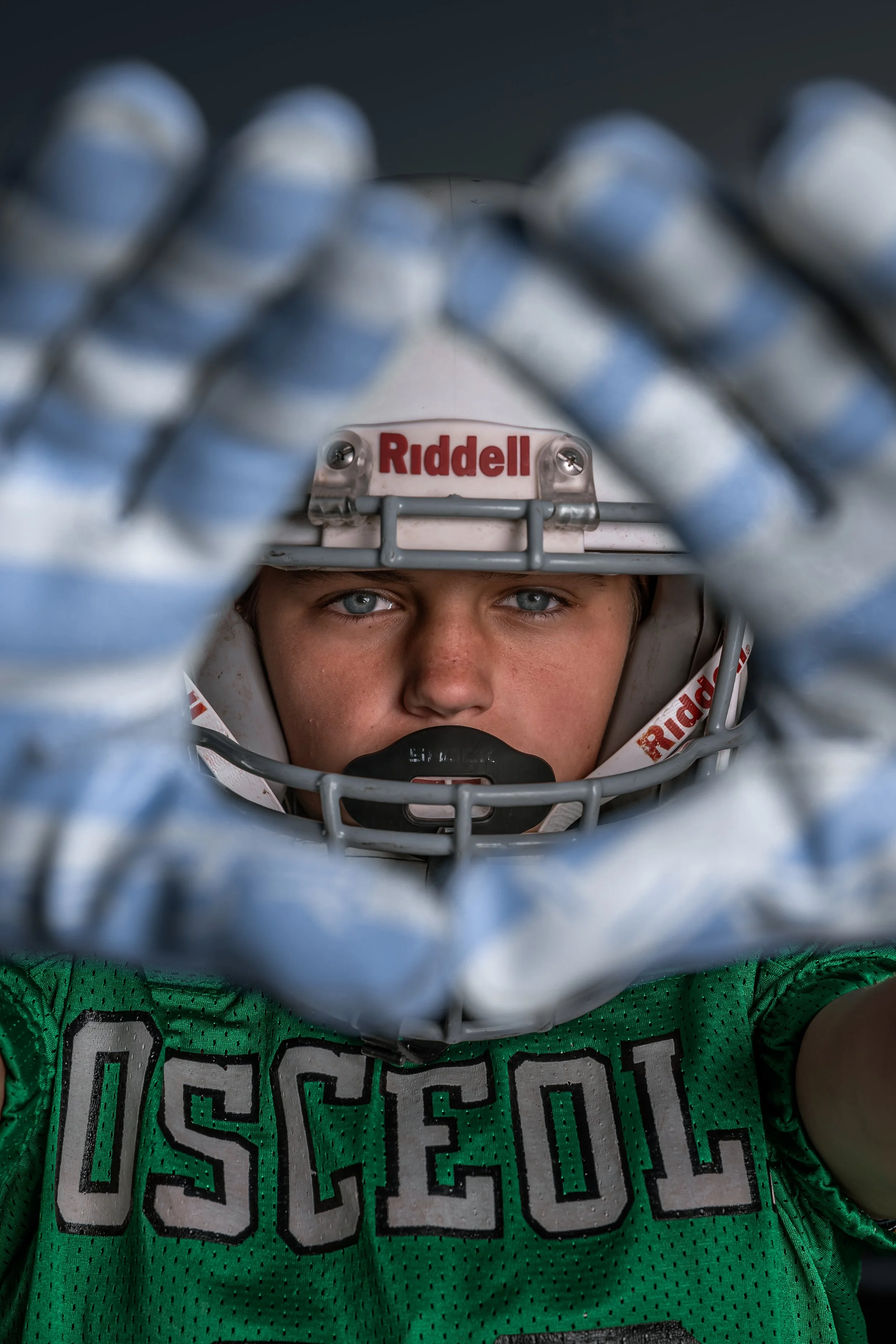 Close-up of a young athlete wearing a green sports jersey and a helmet, seen through a baseball catcher's face mask with blue and white padding, with the brand name 'Riddell' visible on the helmet.