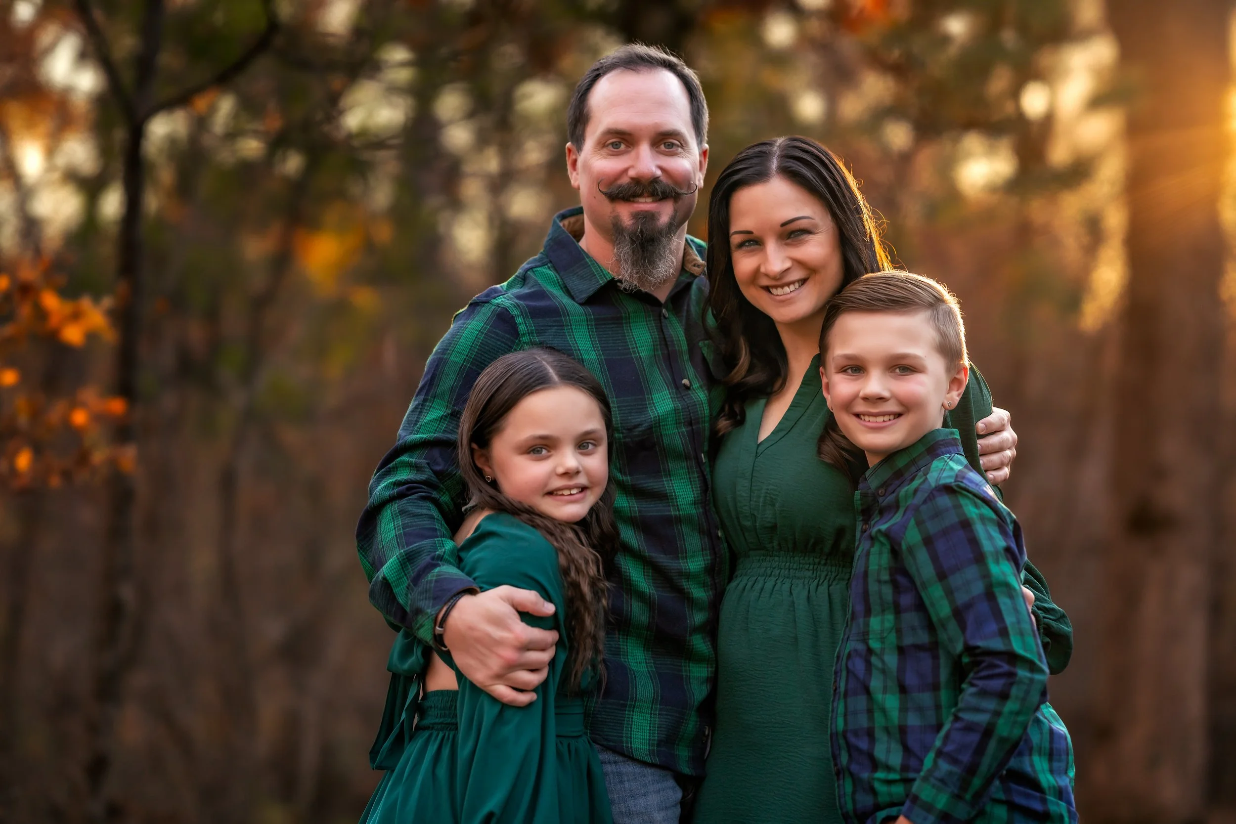 A family of four outdoors during autumn, smiling and embracing each other. The background features fall-colored trees and the setting sun.