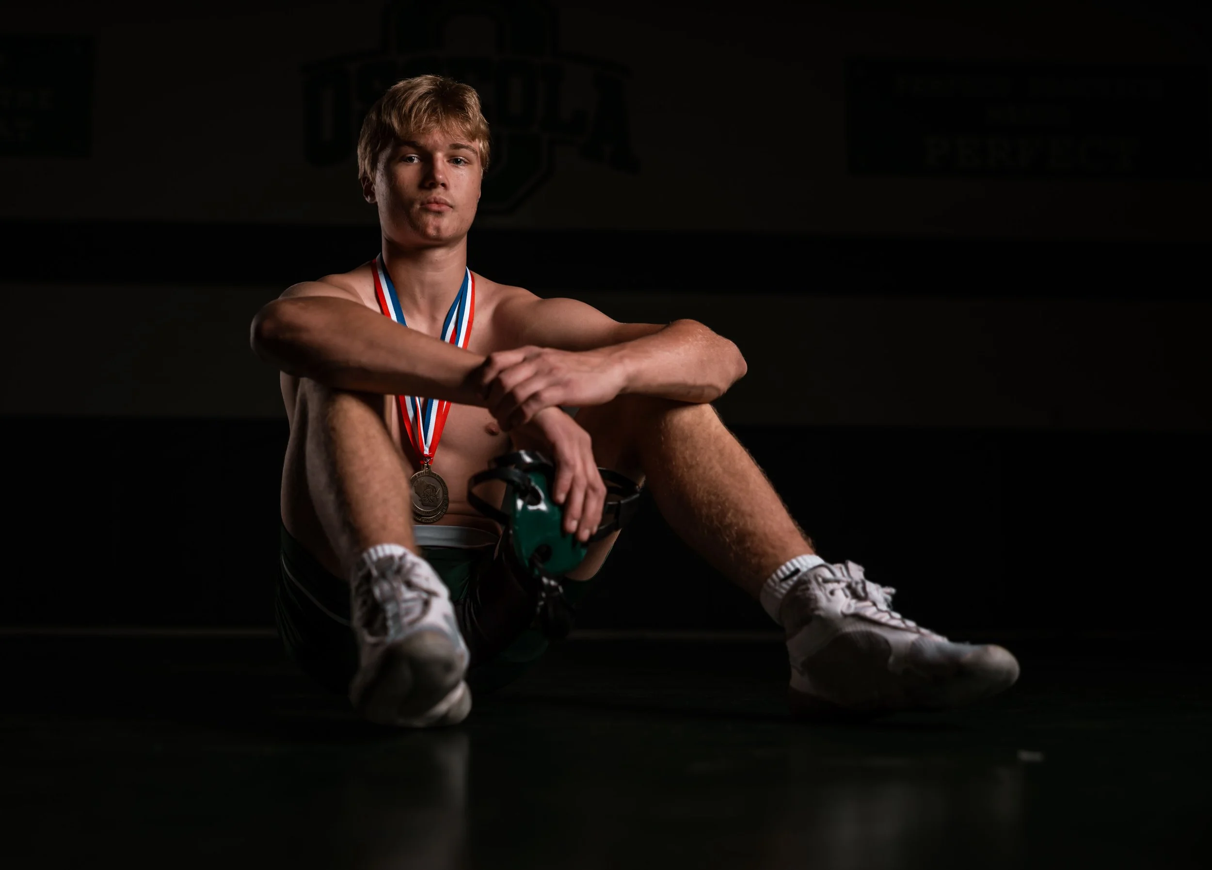 A teenage boy sitting on the floor in a dark gym, wearing athletic shoes and shorts, with medals around his neck, holding a device in his hand, with a serious expression.