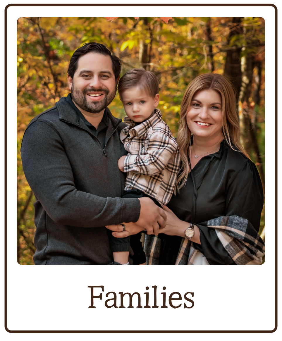 A family of three standing outdoors in autumn, smiling, with a background of colorful fall trees. The father, mother, and young son are dressed in casual, warm clothing.