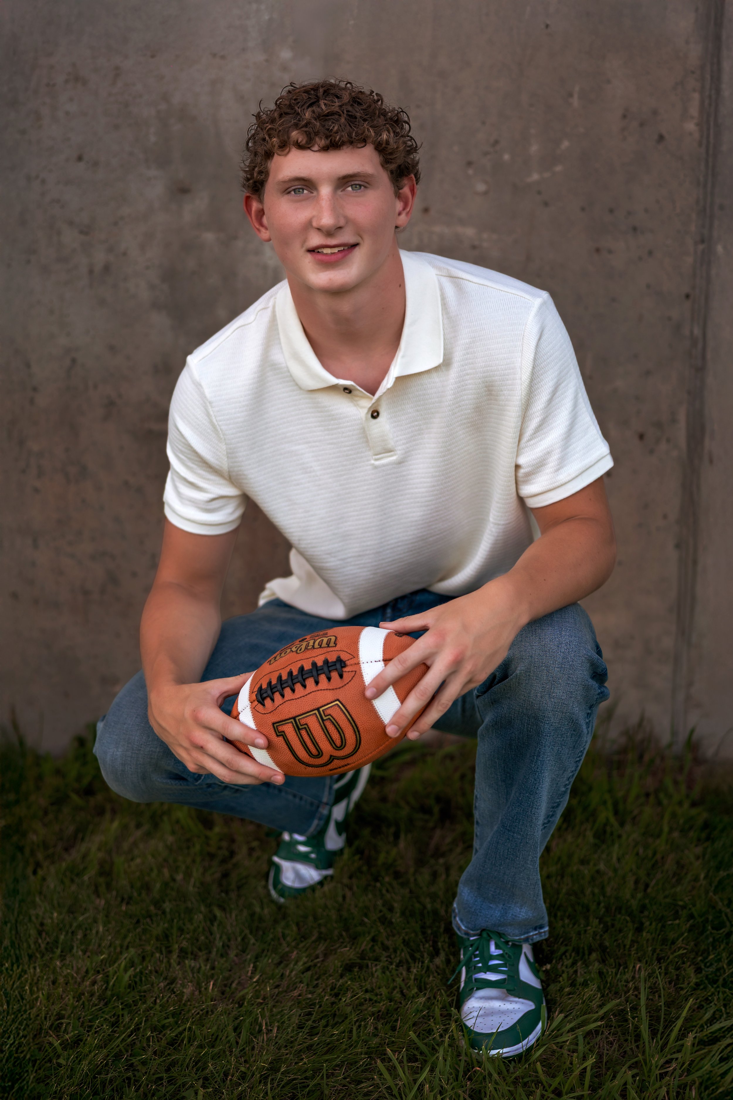 A young man with curly hair, wearing a white polo shirt and jeans, kneeling on grass and holding a football, posing outdoors against a concrete wall.