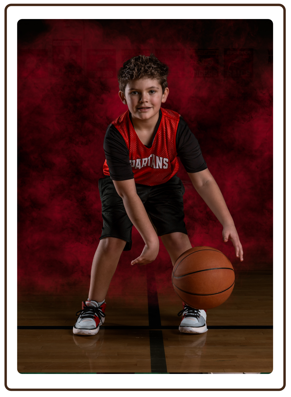 Young boy in a black and red basketball uniform with 'Spartans' lettering dribbling a basketball on a basketball court with a red smoky backdrop.