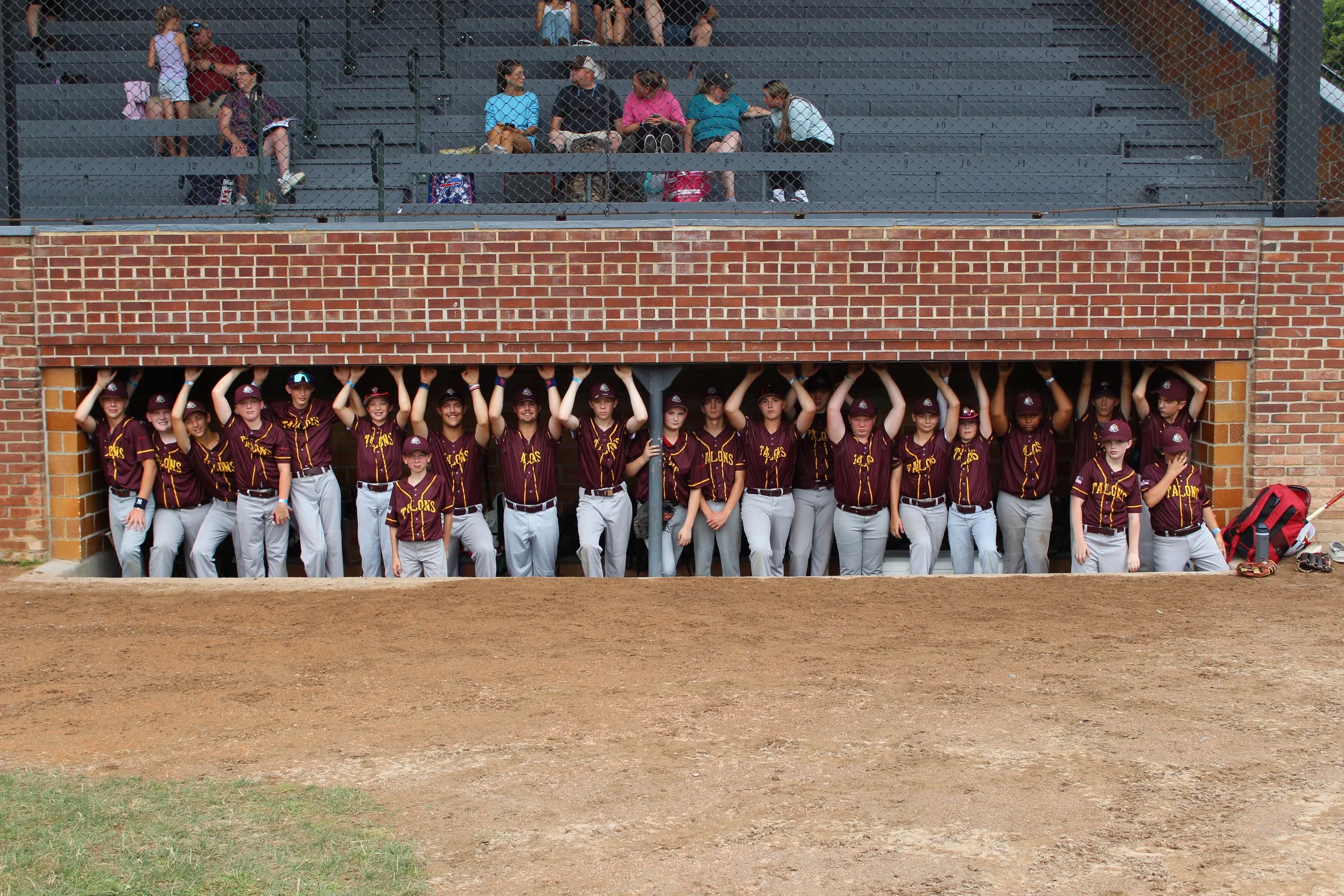 A group of young baseball players in maroon and white uniforms standing beneath a brick dugout, with some raising their hands and others looking towards the camera, with spectators sitting in the bleachers above.
