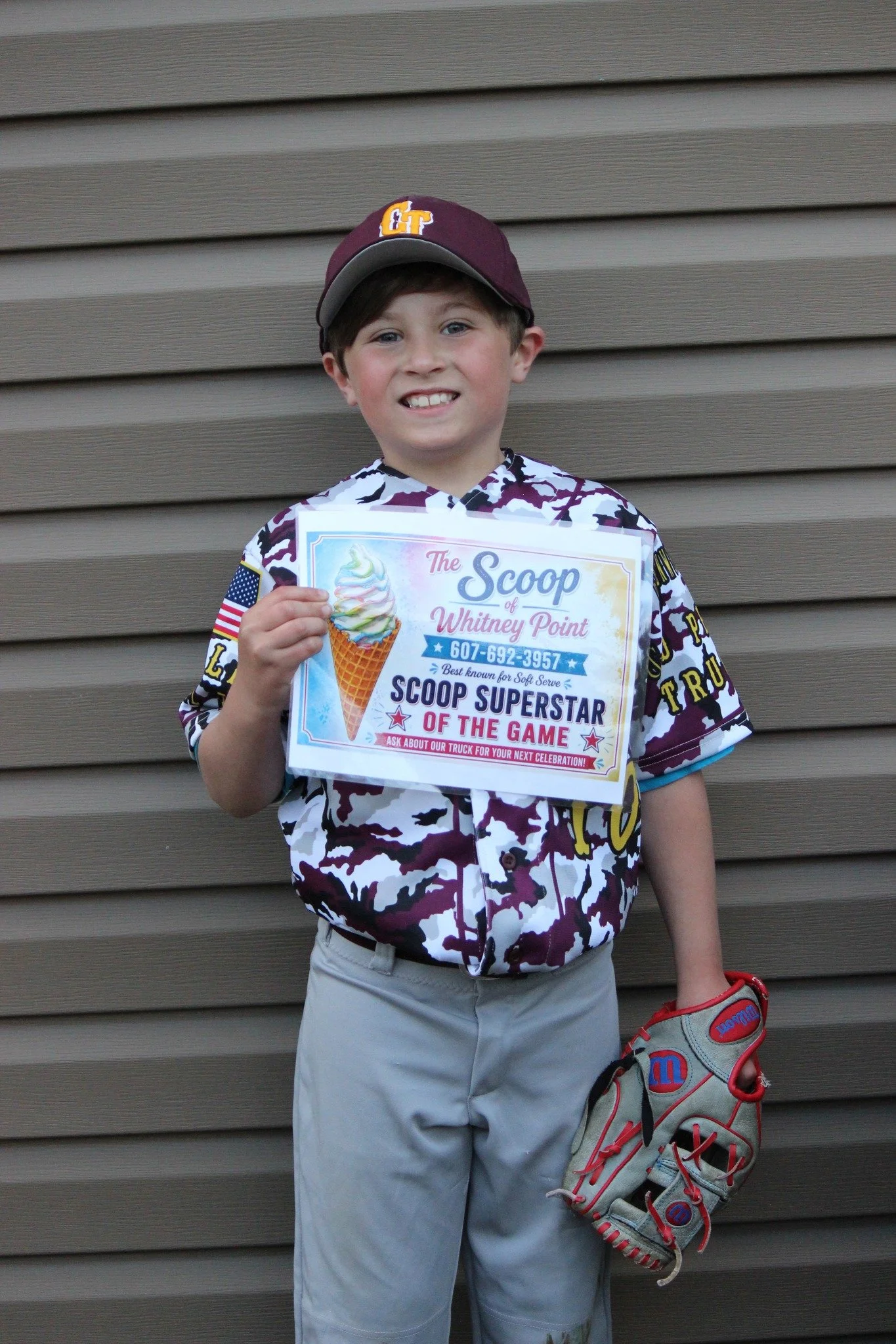 Young boy wearing a maroon and gray baseball cap, holding a sign that reads 'The Scoop of Whitney Point,' standing against a brown wooden wall and holding a baseball glove and shoe.