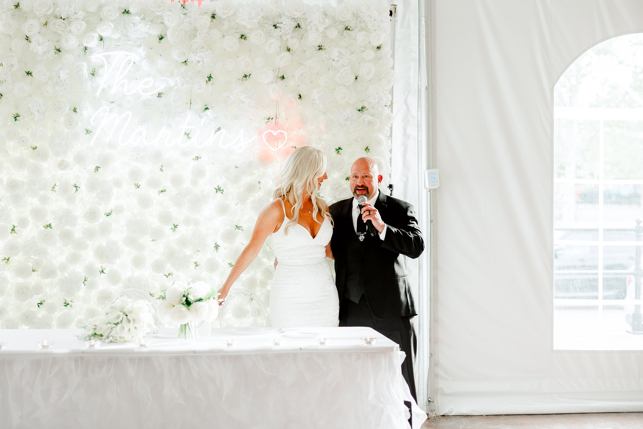 There's a bride and groom, she's looking at him and he's looking out while speaking. they have a beautiful white flower backdrop behind them. He's wearing a black suit and she's wearing a white wedding dress. there's a long table in front of them.