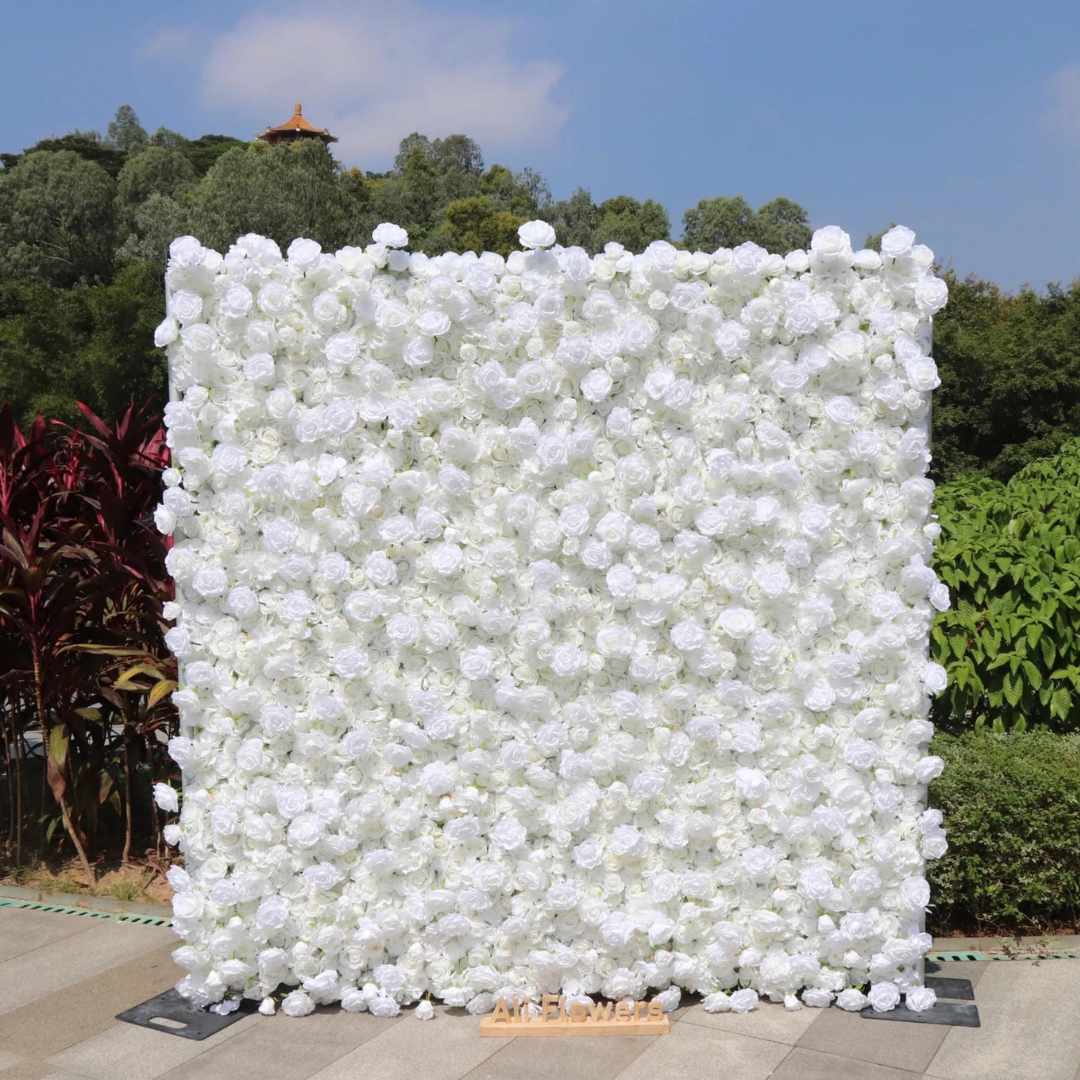 A large square wall of white roses at an outdoor event, with a small wooden sign that reads 'All Flowers' at the bottom, and trees and a small pagoda-style structure in the background.