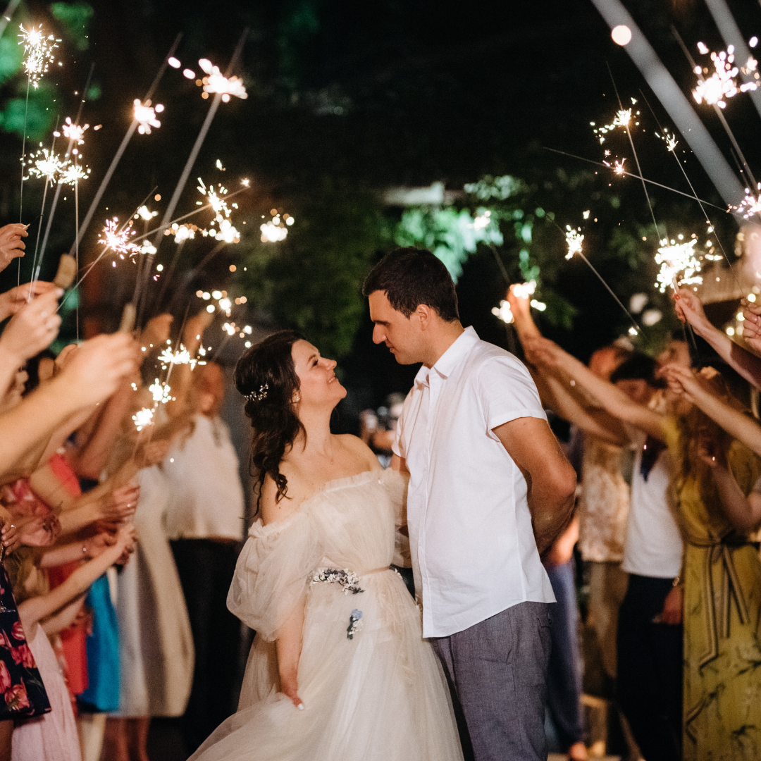 a bride and groom in the middle, looking into each other's eyes. they are doing a beautiful sparkler sendoff. they are both smiling and happy. the background is blurred but it's focused on the couple